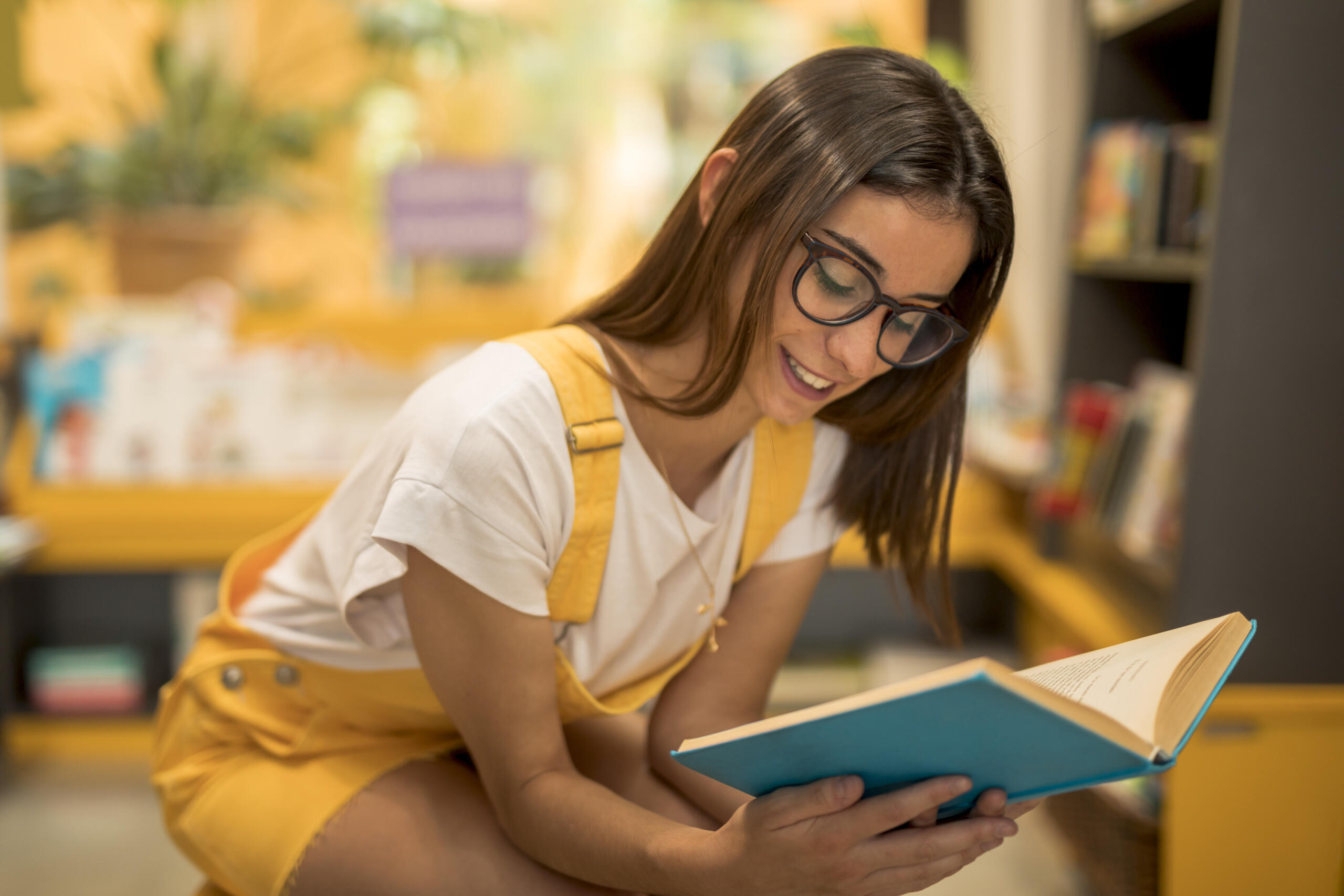 chica sentada con el torso inclinado leyendo un libro en medio de una biblioteca