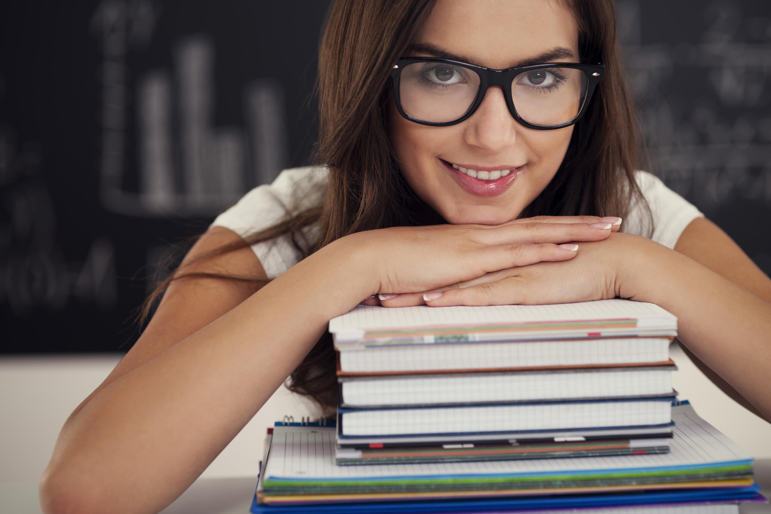 chica con lentes sonriente mirando a la cámara con las manos y el mentón apoyados en una pila de libros y cuadernos