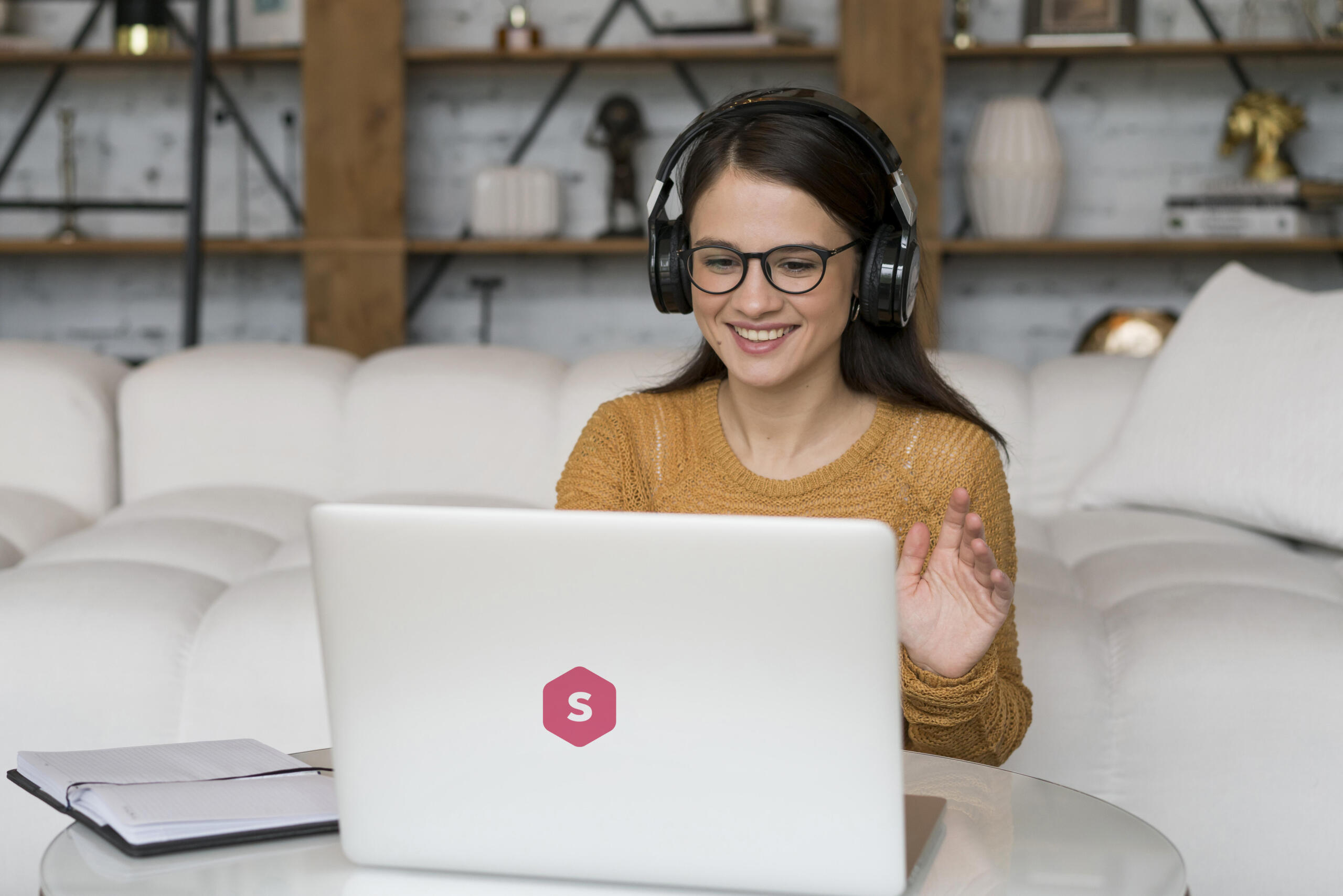 vista de una laptop con sticker del logo Superprof y una chica con lentes sentada en un sofa con audifonos saludando a la pantalla