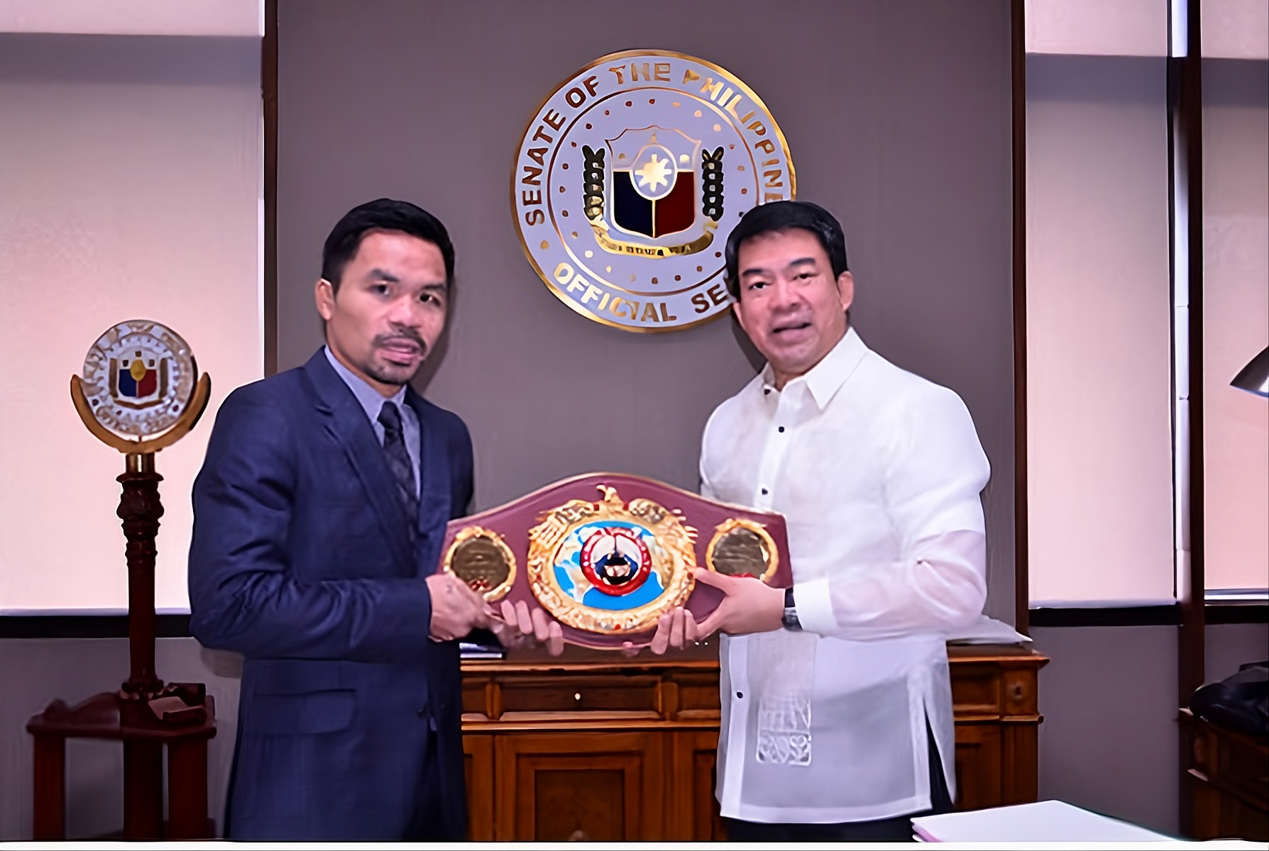 Dos hombres vestidos de gala sosteniendo un cinturón de campeón de boxeo frente al escudo del Senado de Filipinas.