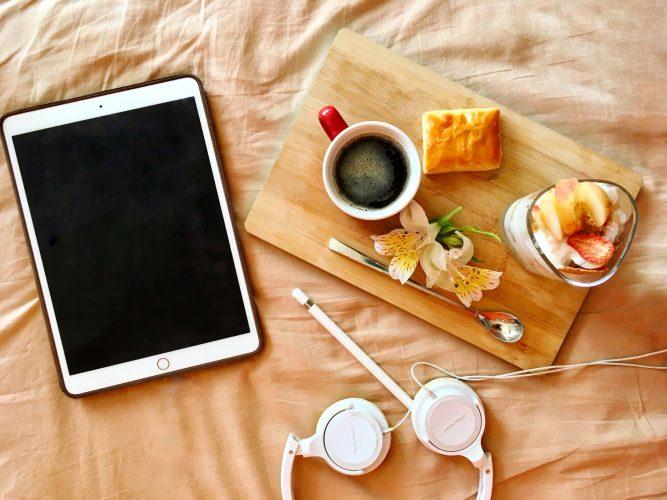 Tablet, białe słuchawki, długopis na pomarańczowym prześcieradle, kubek kawy, ciastko, łyżeczka, kwiat jogurt z owocami w szklance na drewnianej desce.