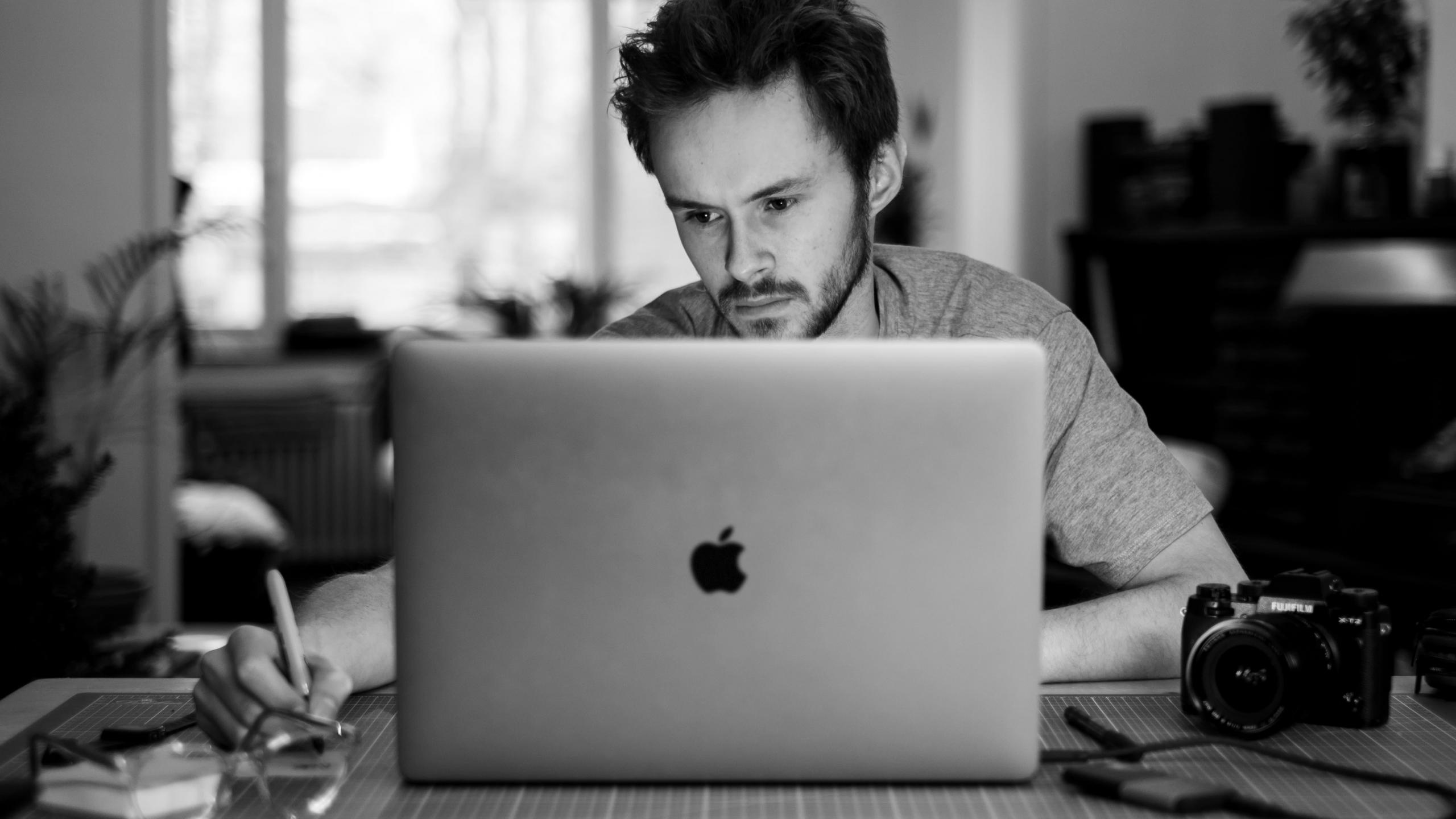 mężczyzna siedzący przed laptopem obok niego na biurku leży aparat fotograficzny