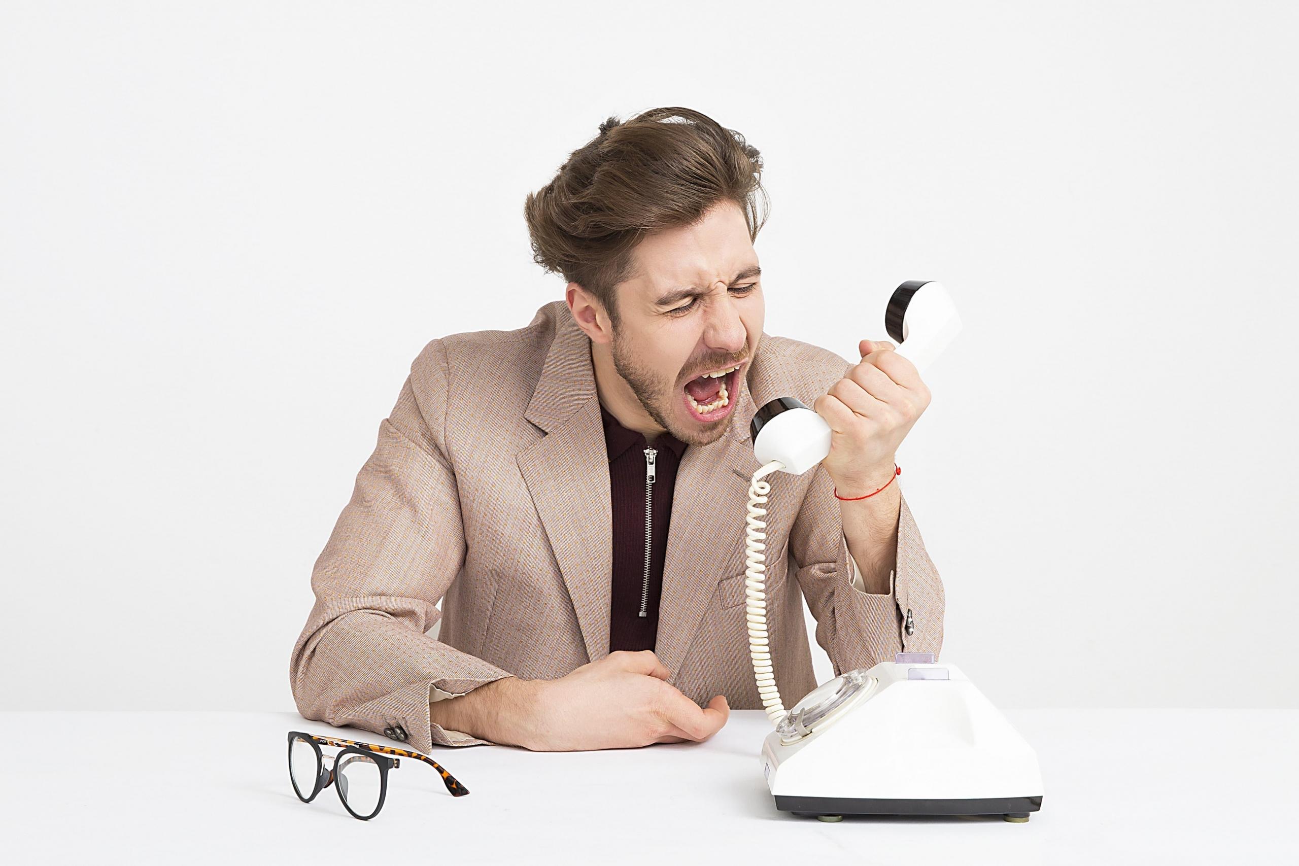 Mężczyzna krzyczący do słuchawki od telefonu analogowego, okulary na białym stole.