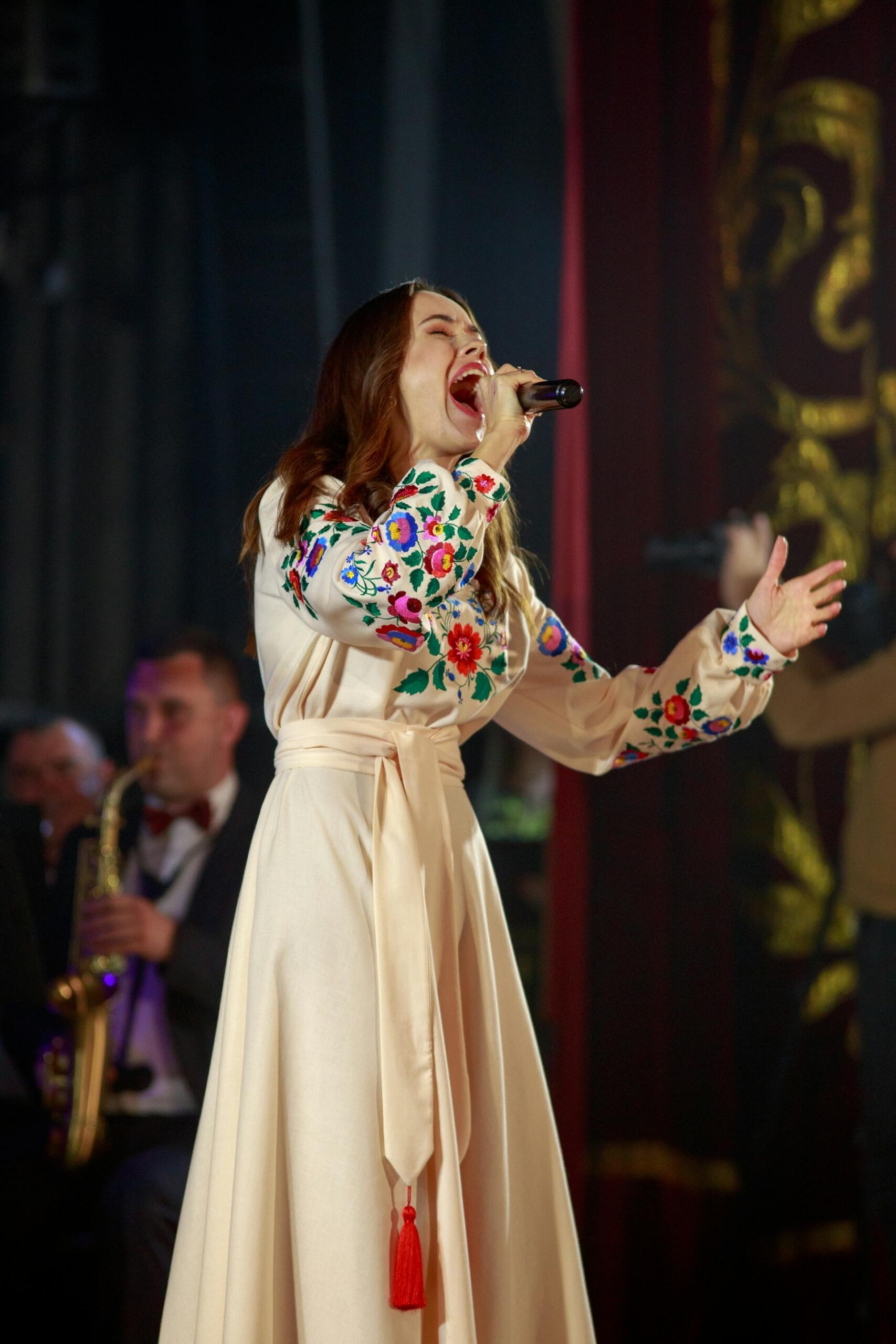Piosenkarka w białej sukience z kwiatowym haftem śpiewa do mikrofonu na scenie, a za nią w tle widać zamazaną postać saksofonisty.