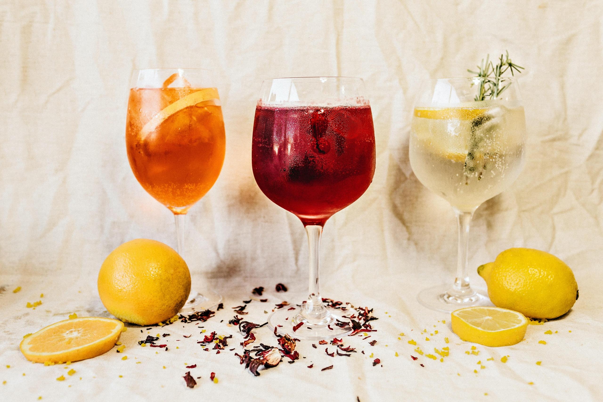 Trzy kieliszki od wina z różnokolorowym alkoholem, na białym obrusie, cytrusy, cytryna, pomarańcza.