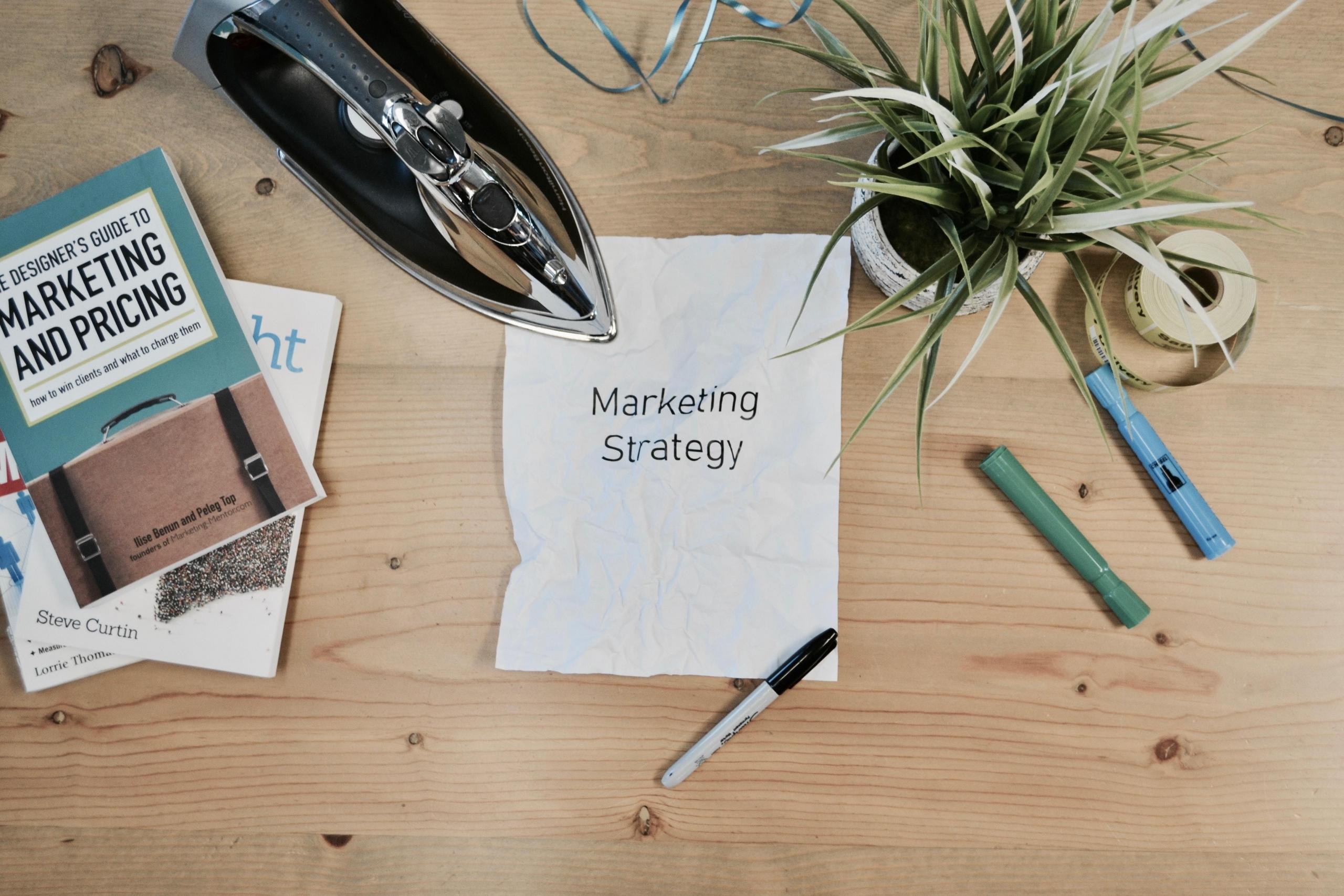 Drewniany blat z przyborami do pisania, kwiatem doniczkowym i żelazkiem, na środku kartka z napisem "Marketing Strategy".
