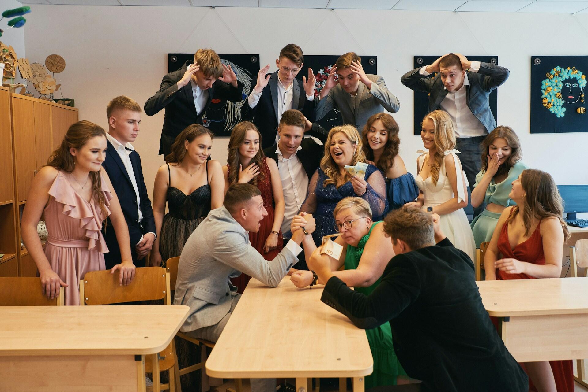 Uczniowie ubrani na galowo w klasie, gdzie mężczyzna siłuje się na rękę z nauczycielką przy wiwacie.