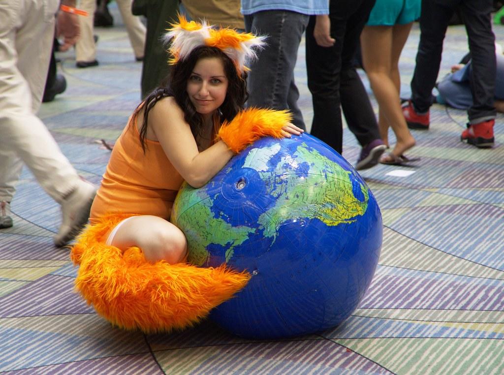 Kobieta cosplayerka w kostiumie lisa Mozilla Firefox z piłką z wizerunkiem świata, na podłodze, wśród tłumu ludzi.