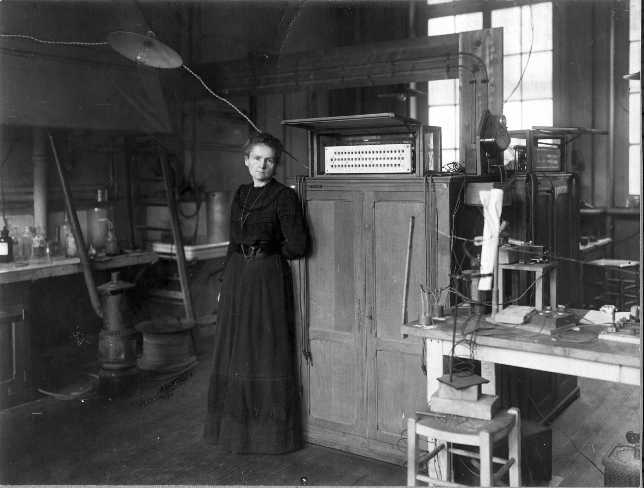 Maria Skłodowska-Curie w laboratorium, sprzęt chemiczny, szklane pojemniki, stoły, krzesło.