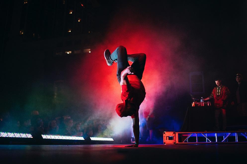 Mężczyzna w czerwonej bluzie tańczący breakdance na scenie w klubie w otoczeniu dyskotekowych świateł do muzyki puszczanej przez DJa.