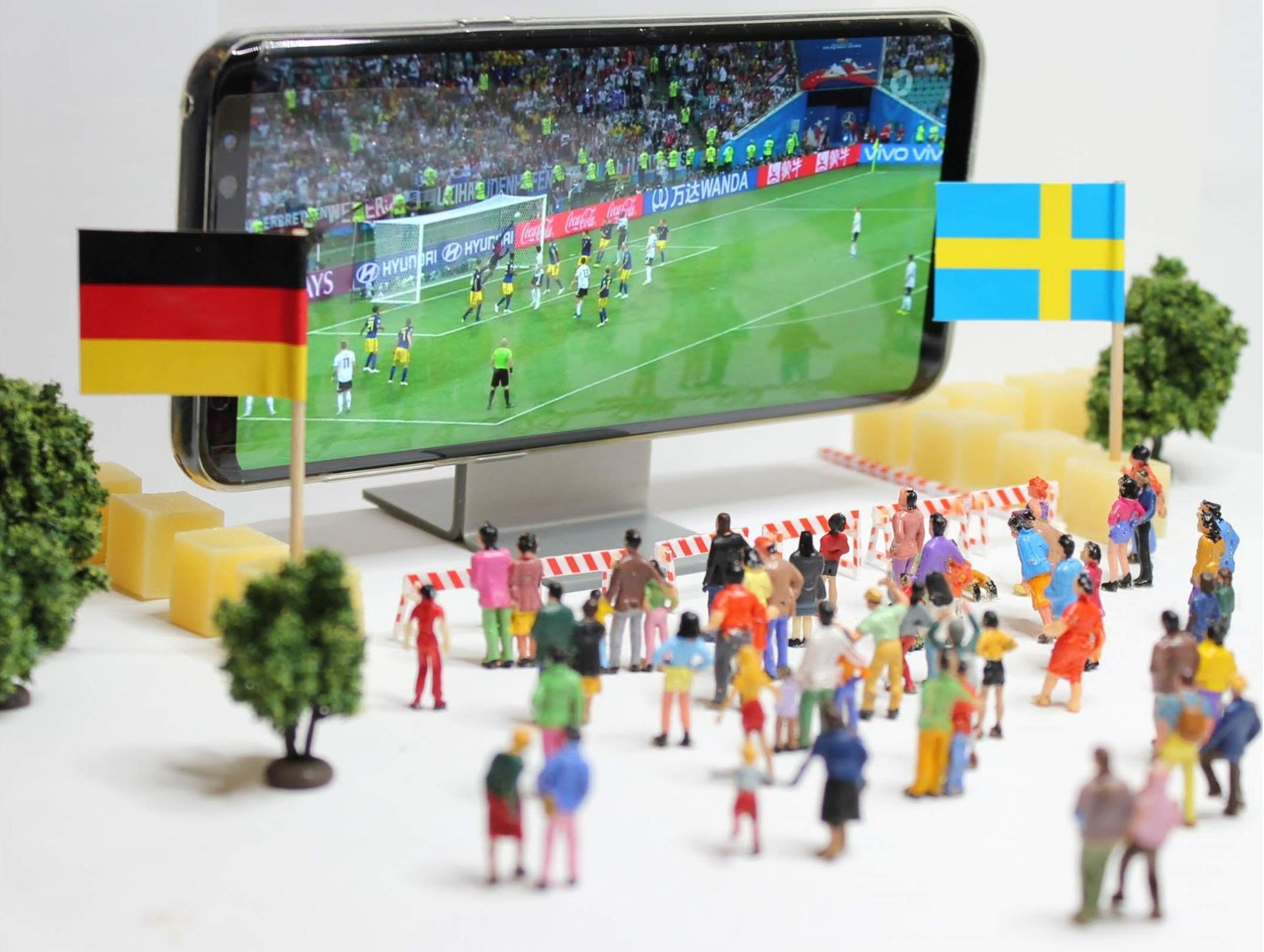 figurki ludzi z flagami oglądających mech niemiec ze szwecją na smartfonie imitującym telebim