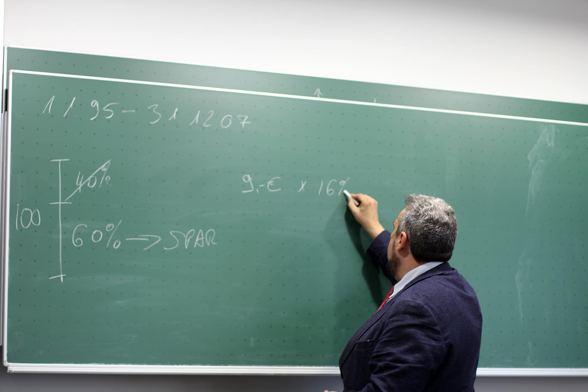 starszy mężczyzna w garniturze rozwiązujący zadanie z matematyki na dużej zielonej tablicy