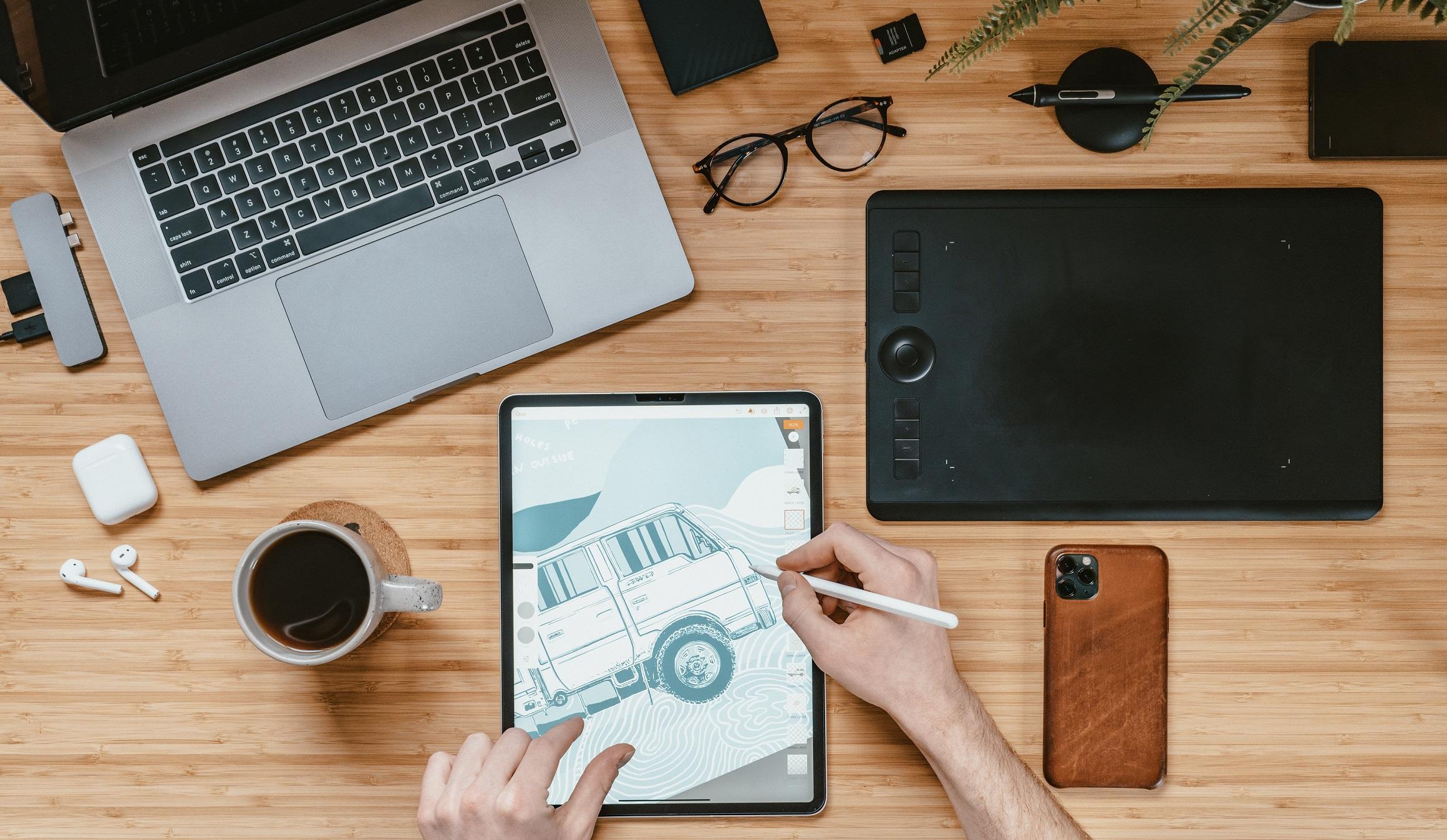 biurko na którym leżą tablet graficzny laptop kubek z kawą smartfon okulary dyski zewnętrzne oraz bezprzewodowe słuchawki i dłoń rysująca na tablecie