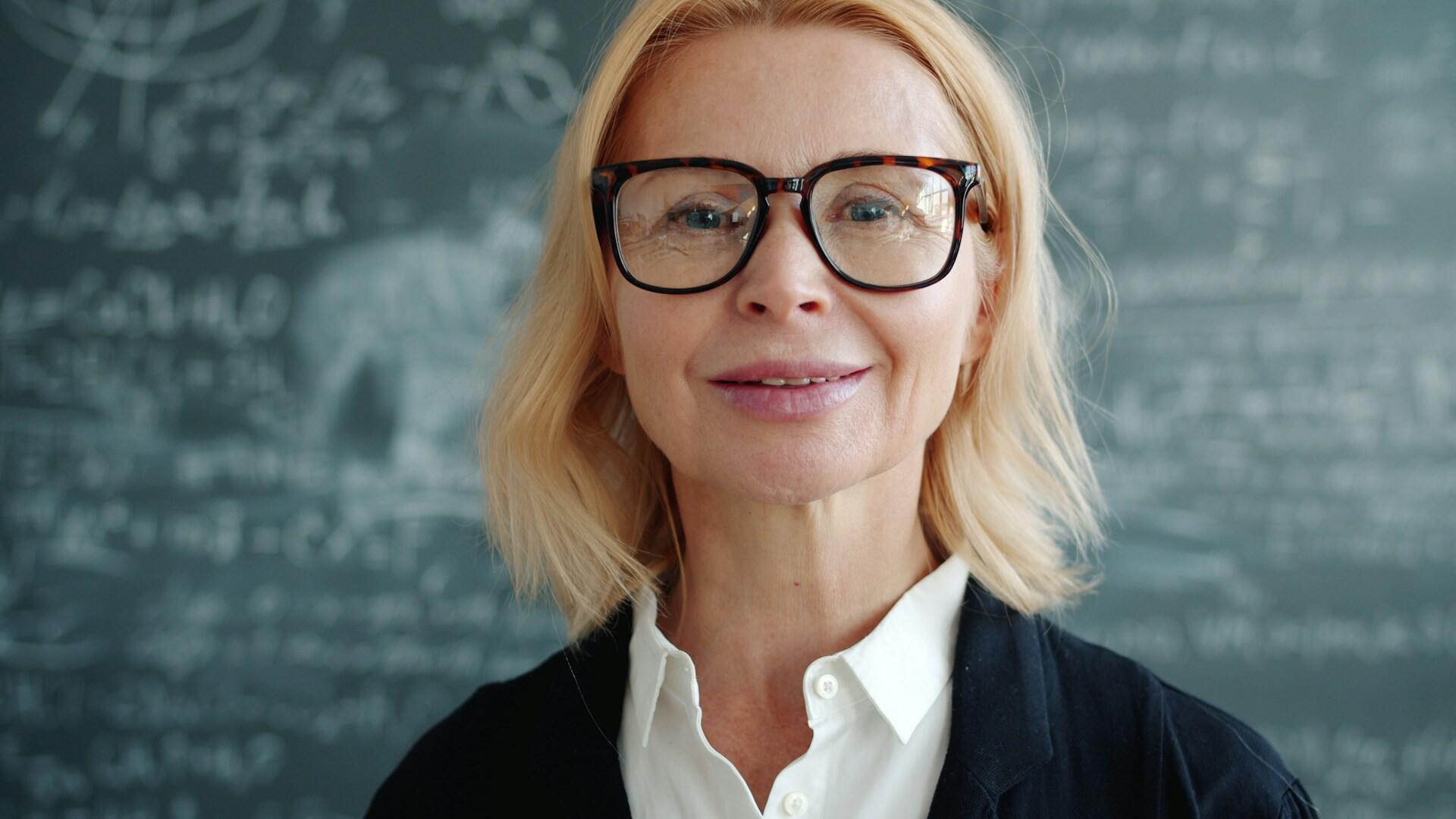 Blondynka w okularach, białej koszuli i ciemnej marynarce stojąca przed tablicą pokrytą równaniami matematycznymi.