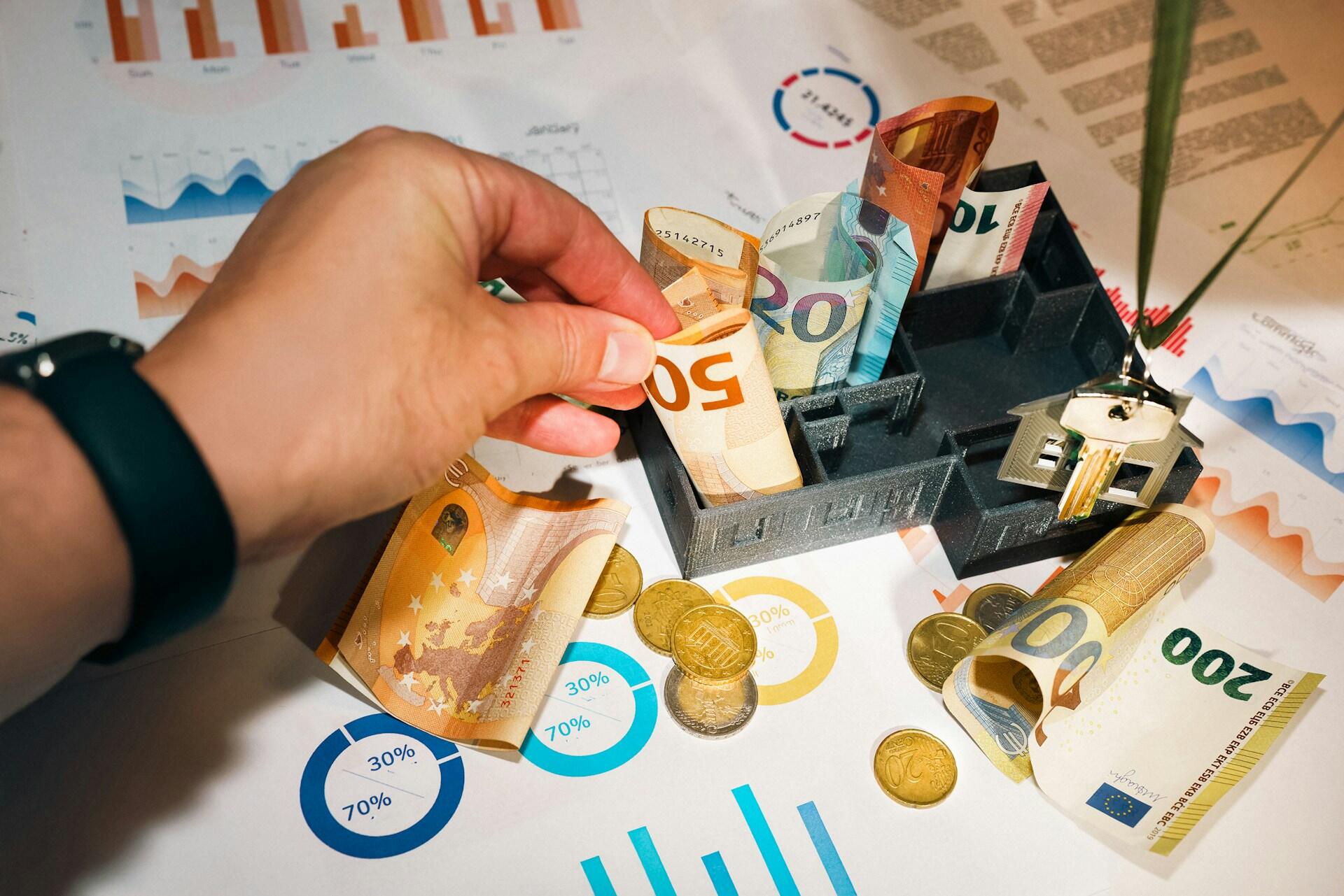 Ręka wkładająca banknoty euro do modelu małego domku, otoczonego monetami, wykresami i kluczami do domu, przedstawionego na dokumencie finansowym.