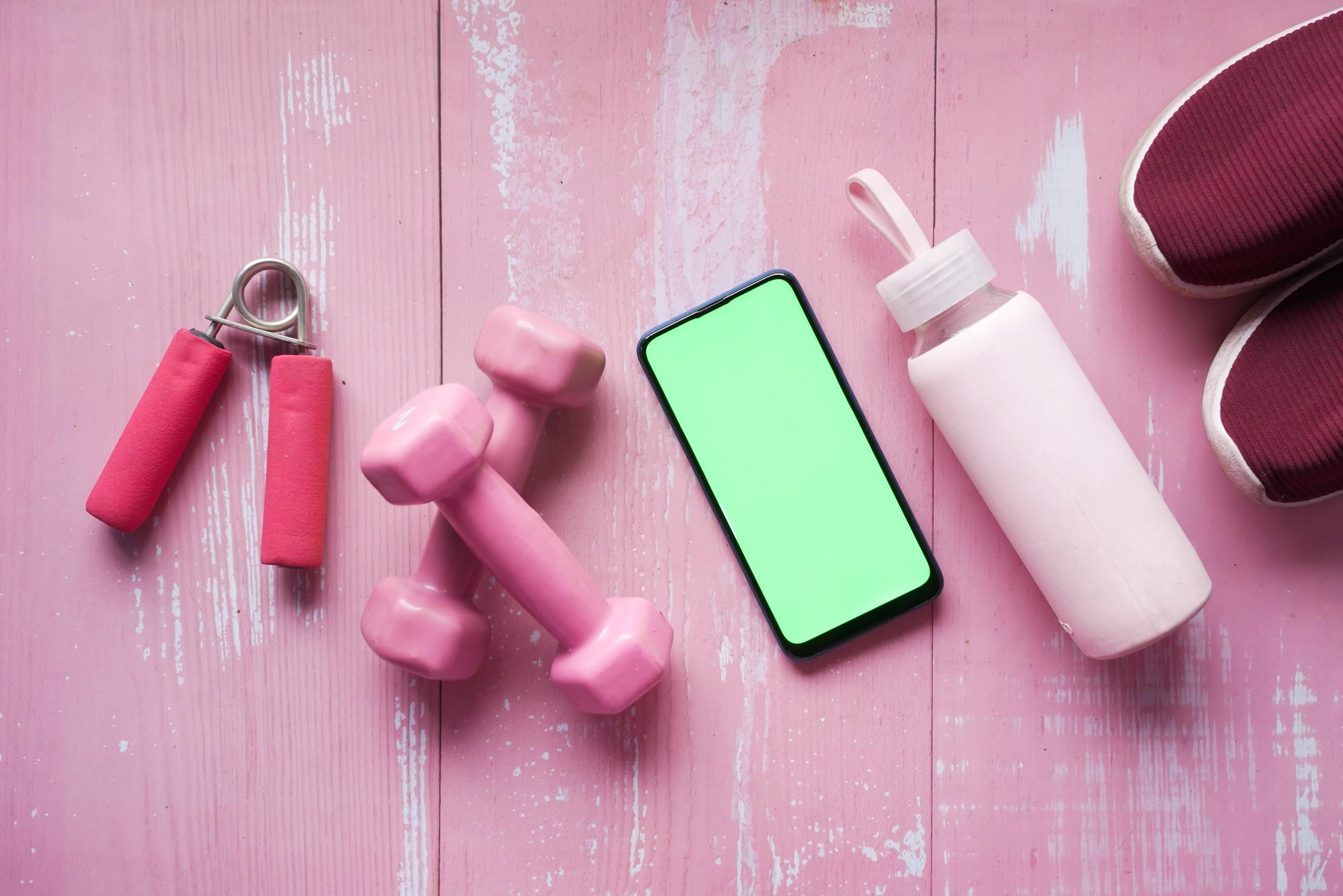 sportowe buty bidon smartfon hantle i ściskacz ręczny leżące na różowej podłodze