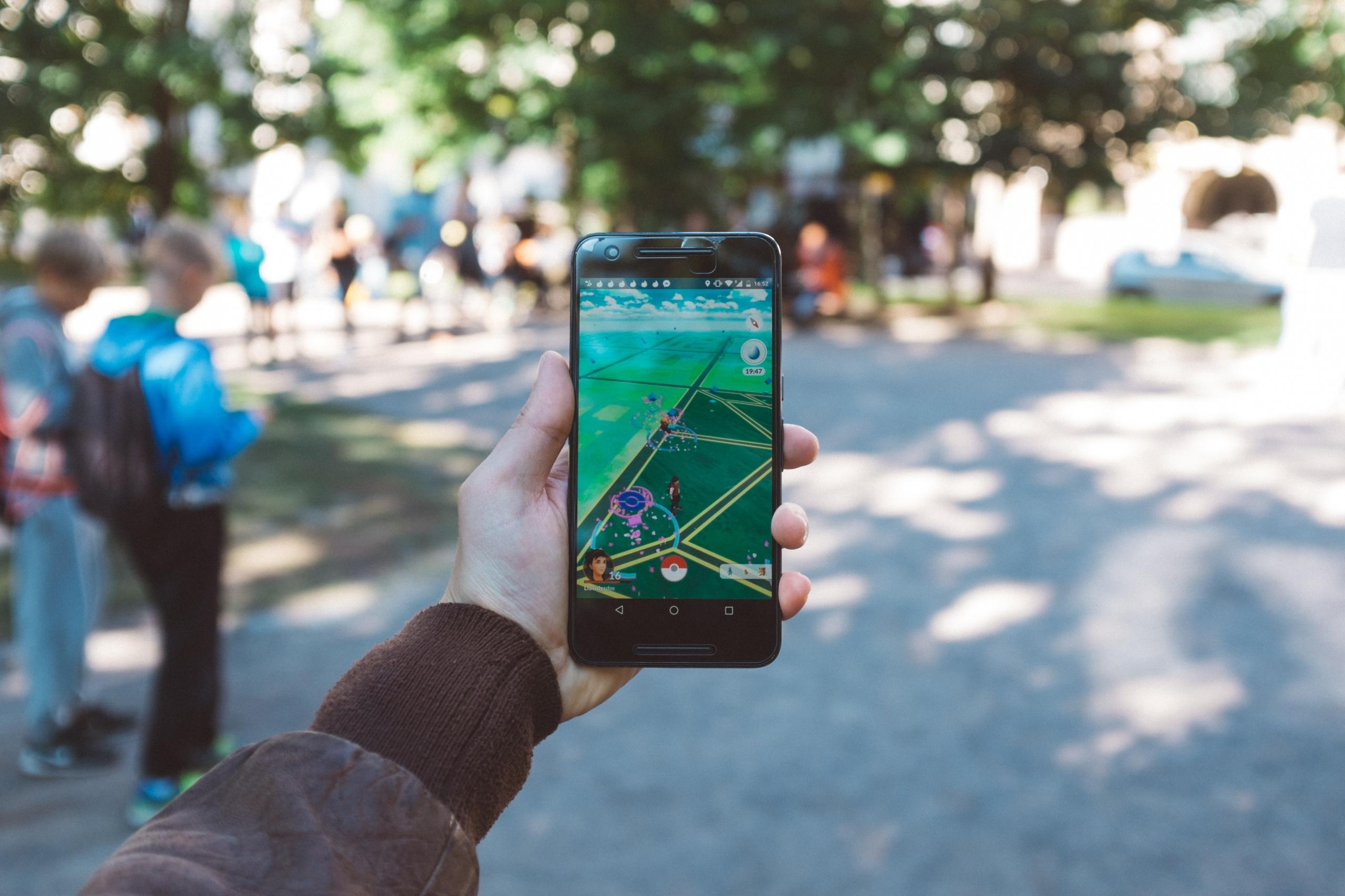 dłoń trzymająca smartfona z włączoną grą pokemon go w tle widać park