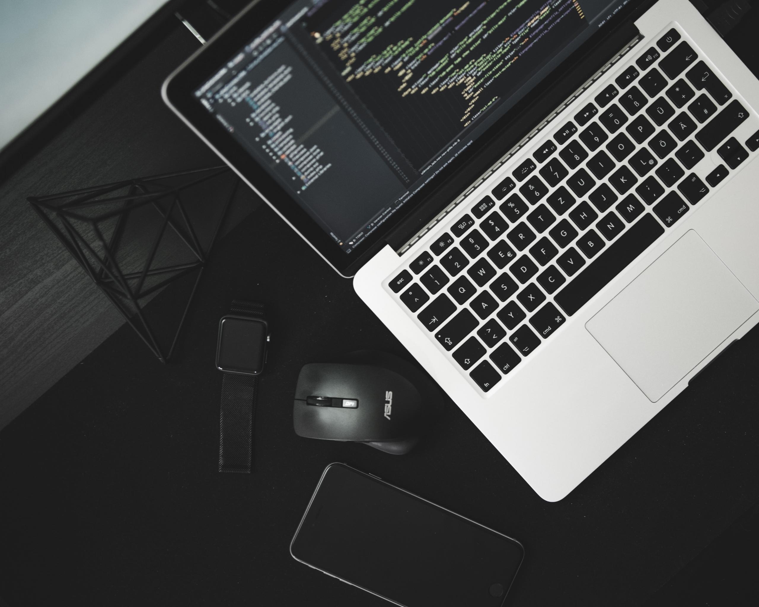 laptop na którym pisany jest program obok leżą na biurku smartfon myszka zegarek i ozdoba
