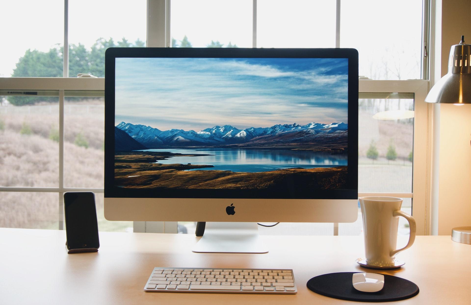 komputer laptop Mac kubek okno drewniane biurko lampka pole las