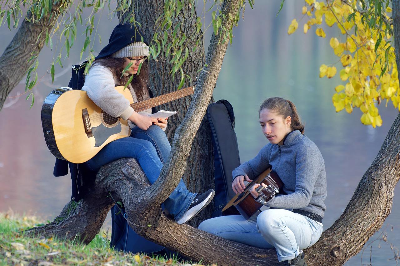 Dwie dziewczyny w parku, siedzące na drzewie i grające na gitarach akustycznych.