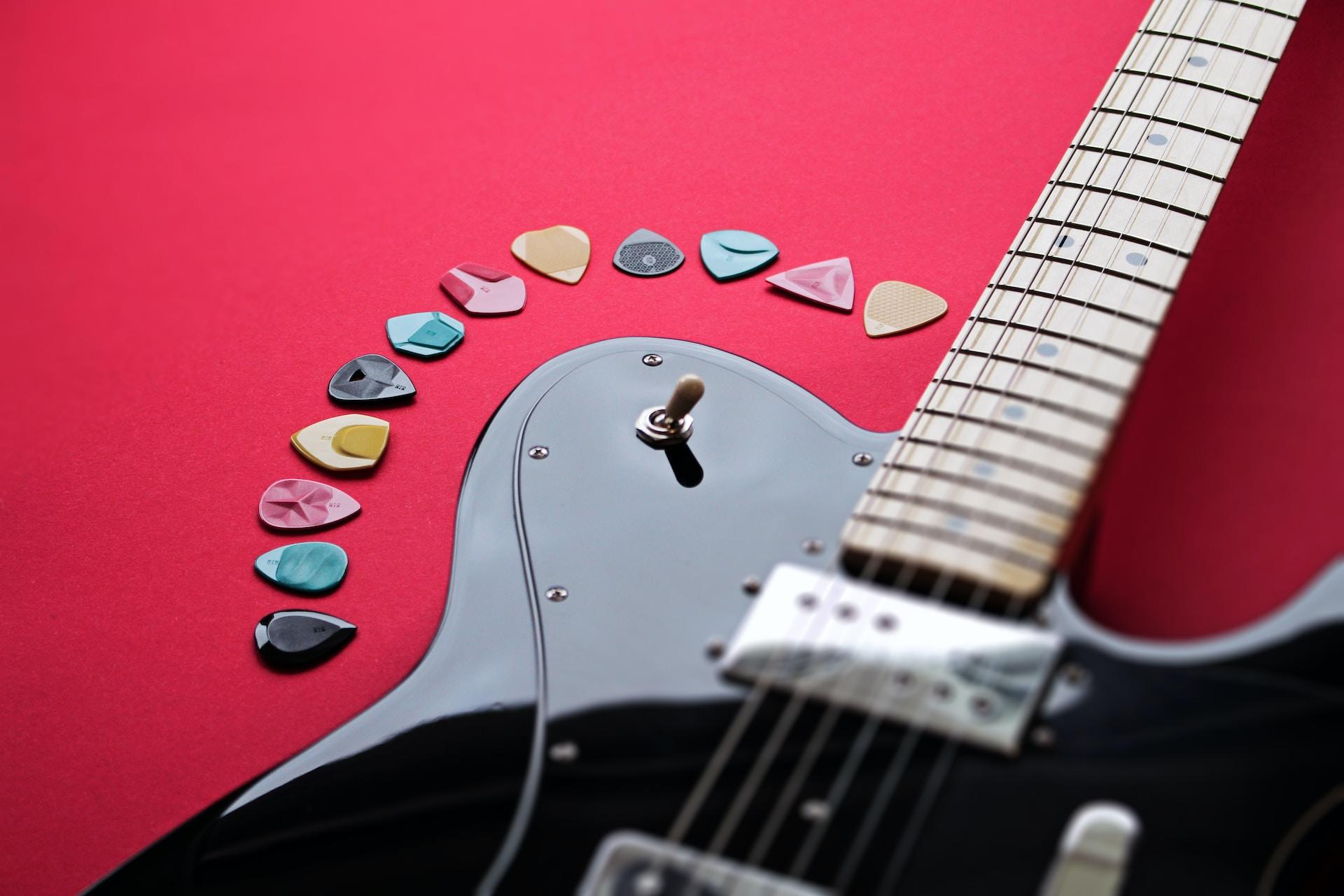 Czarna gitara elektryczna i kolorowe kostki do gry na czerwonym tle.