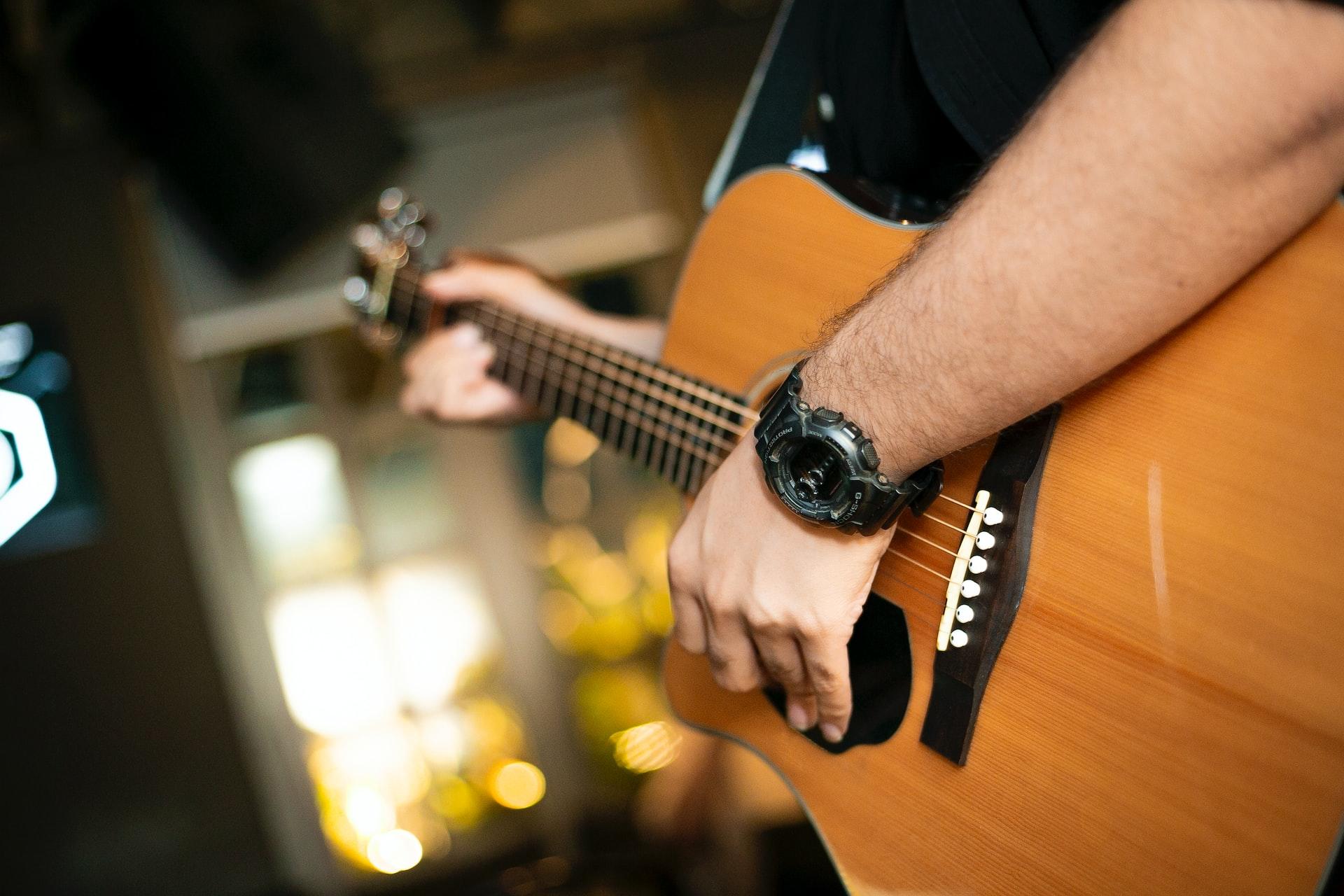 Mężczyzna grający na gitarze akustycznej z zegarkiem na nadgarstku i oknem w tle.