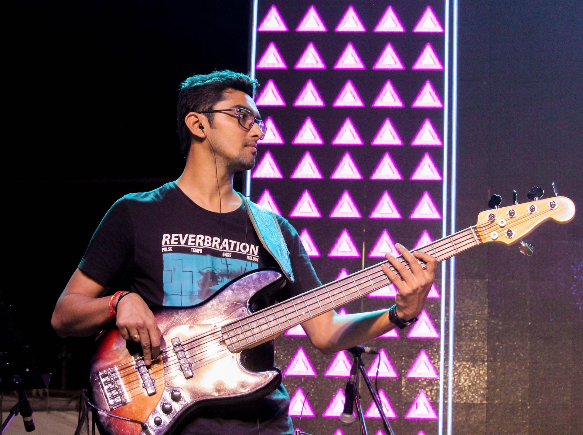 Mężczyzna w okularach, grający na elektrycznej gitarze basowej, na scenie z różowym oświetleniem.