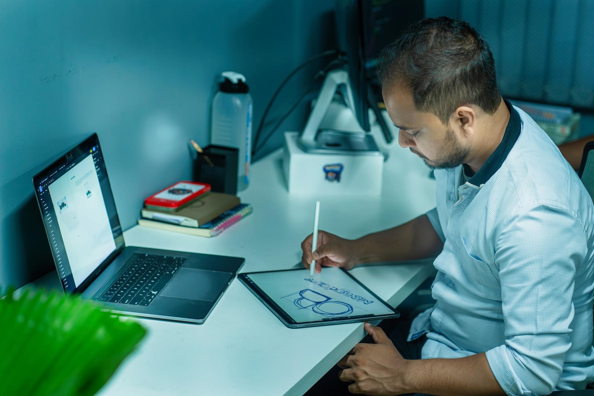 Mężczyzna przy białym biurku, laptop, tablet, lampka i akcesoria biurowe.