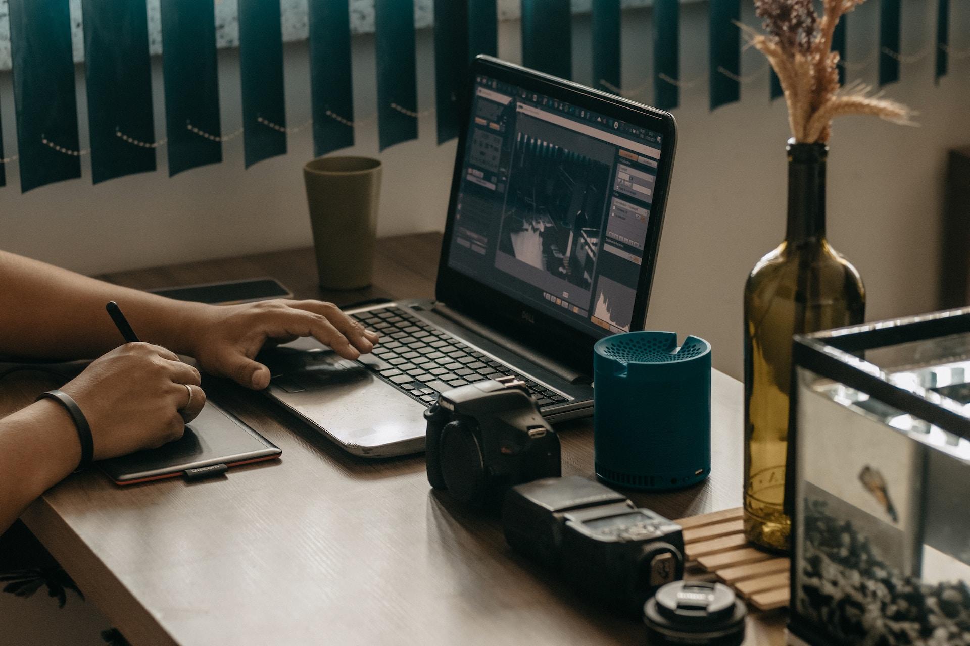 Dłonie mężczyzny na laptopie na drewnianym biurku, wazon, kubek, aparat fotograficzny.