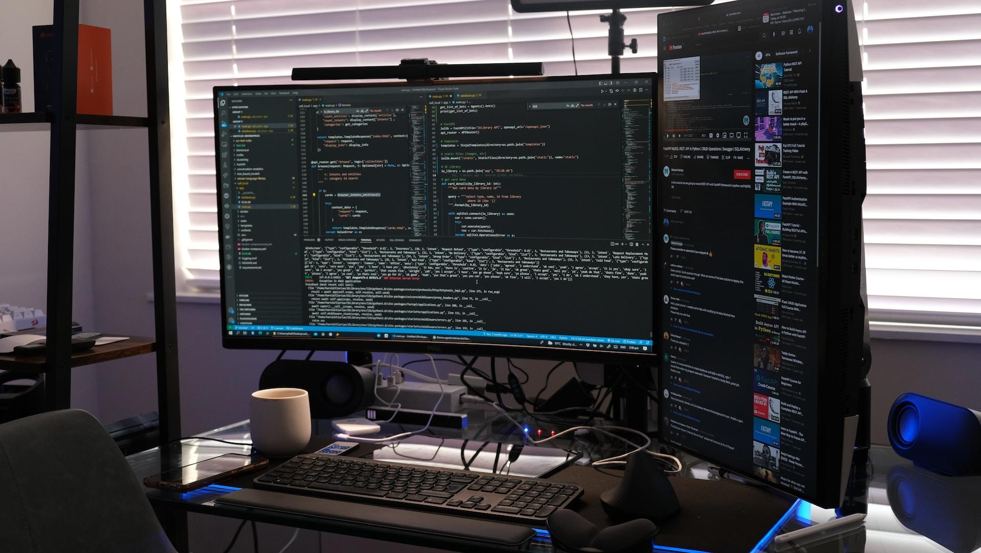 Dwa monitory komputerowe, klawiatura, kubek na biurku, okno zasłonięte roletą, kod programowania.