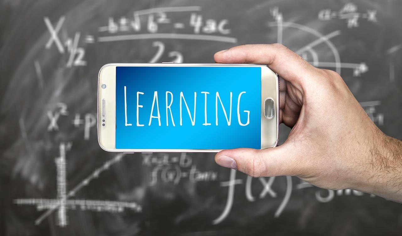 dłoń trzymająca telefon, na którym jest niebieskie tło i napis ''learning'', w tle obrazka tablica z wzorami matematycznymi