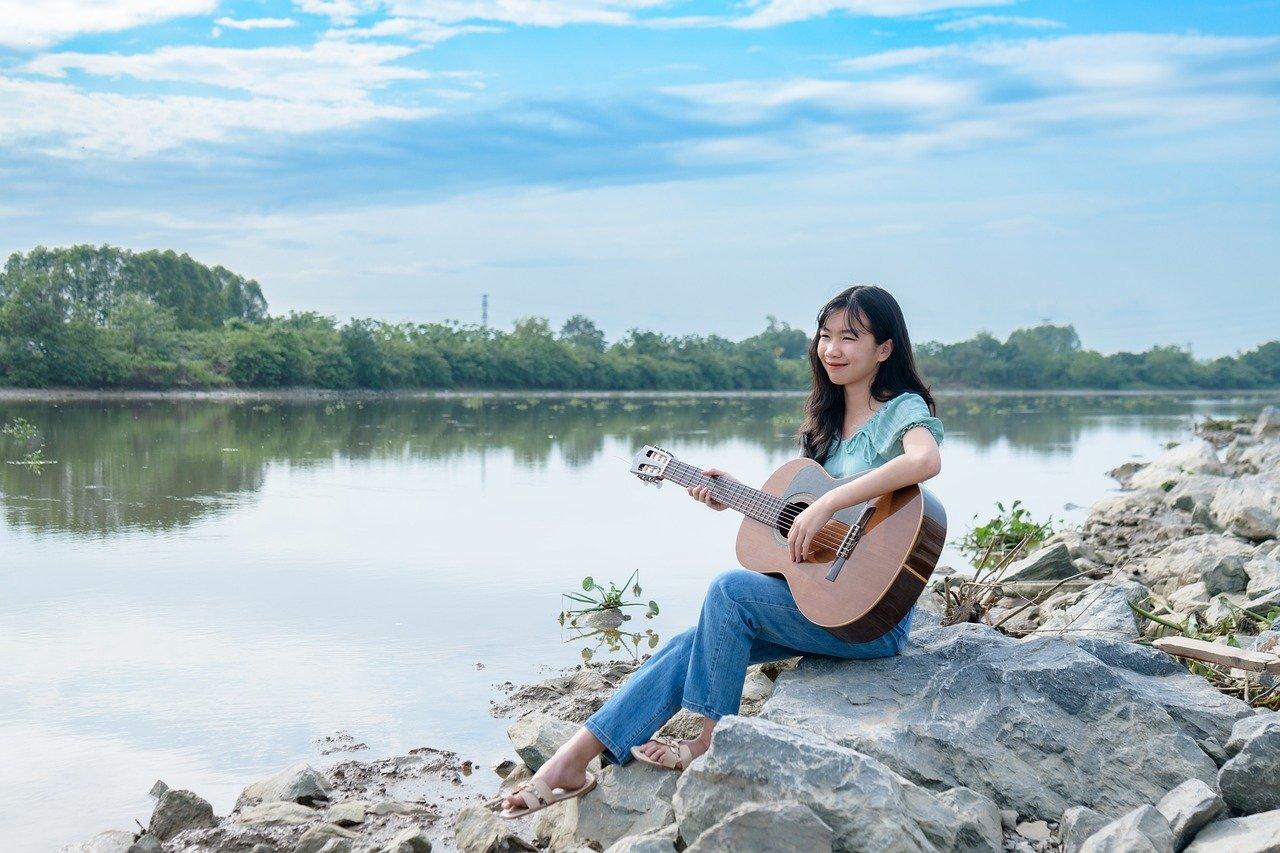 Kobieta z gitarą na plaży, kamienie, woda, roślinność.