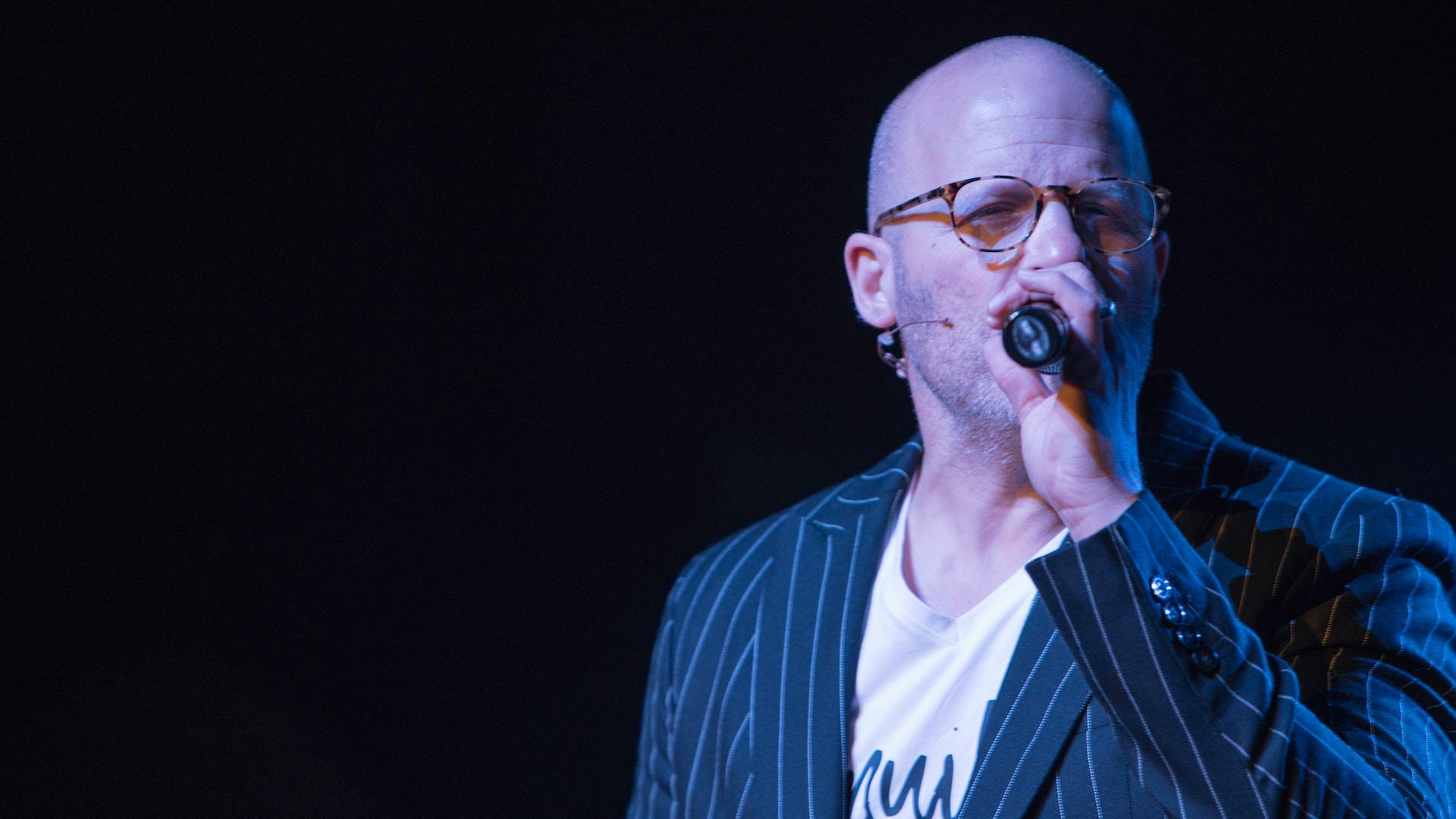 Łysy mężczyzna w garniturze i okularach, śpiewający do mikrofony.