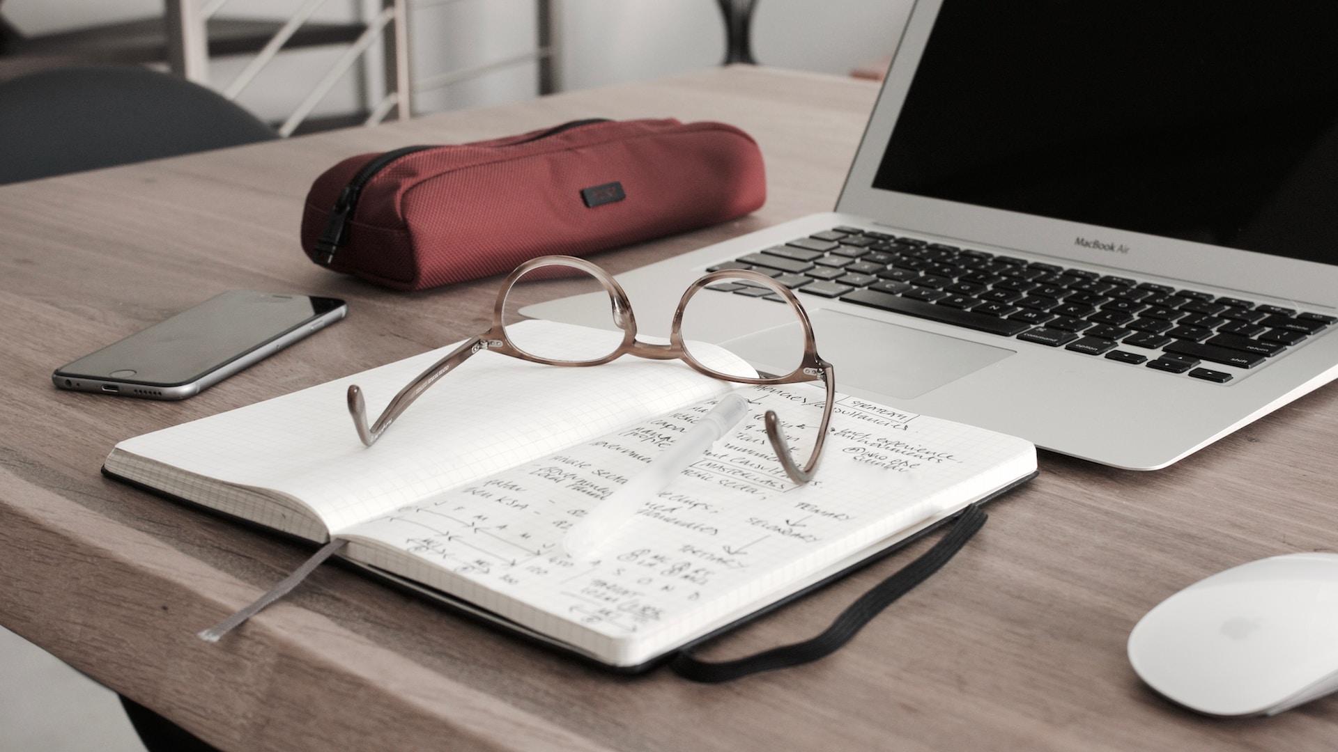 Zeszyt, okulary, laptop, piórnik na biurku.
