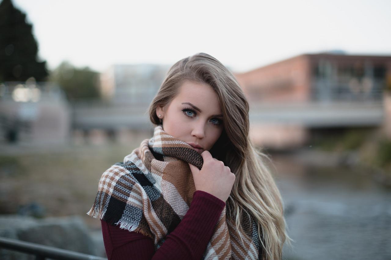na pierwszym planie blond dzieczyna w swetrze bordo i szaliku w kratkę, w tle budynki