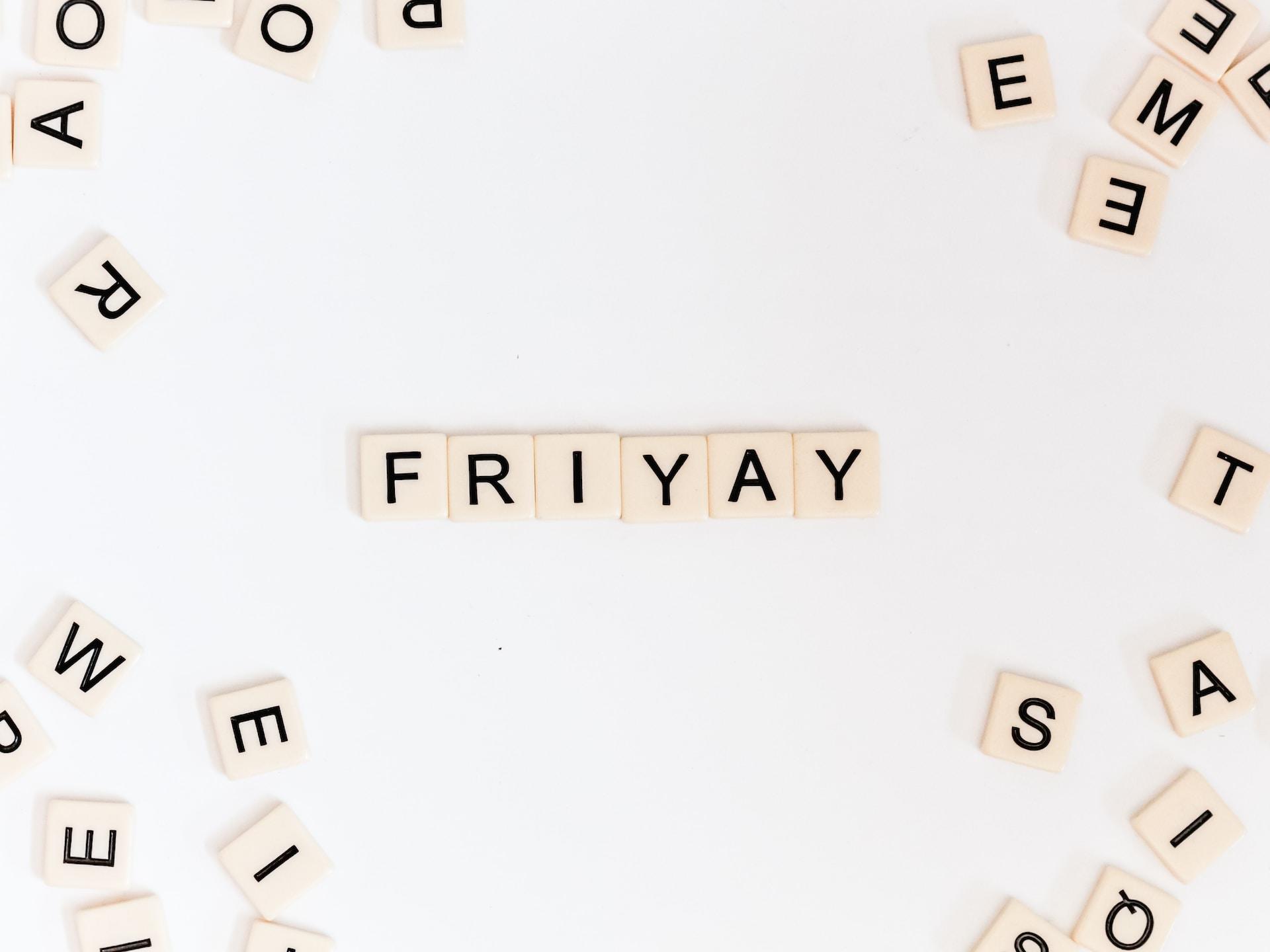 Białe klocki z czarnymi literami na białym stole, ułożone w napis FRIYAY, klocki luzem w rogach.