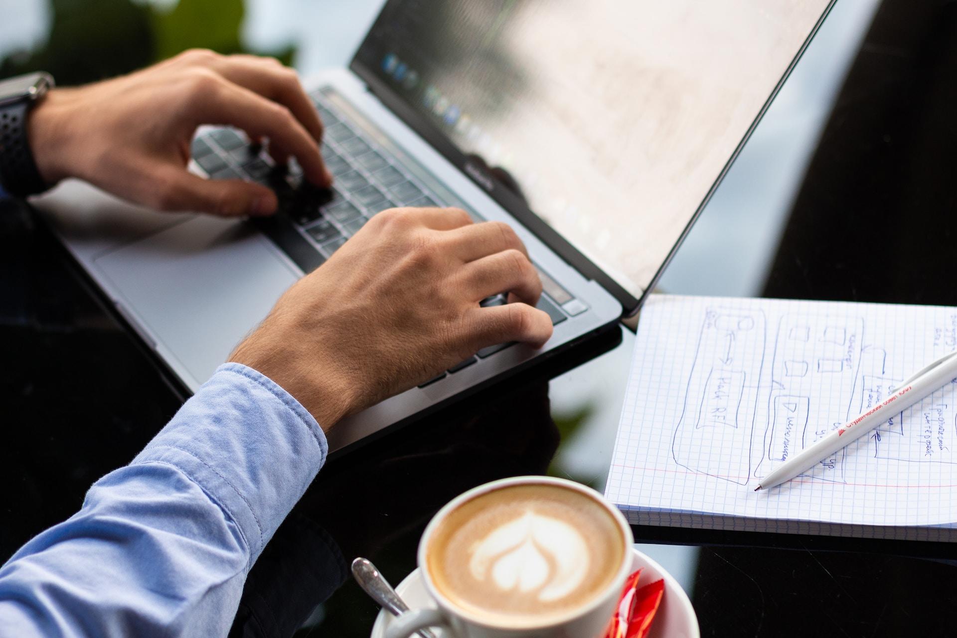 Mężczyzna przy komputerze, laptop, kawa, notes, długopis.