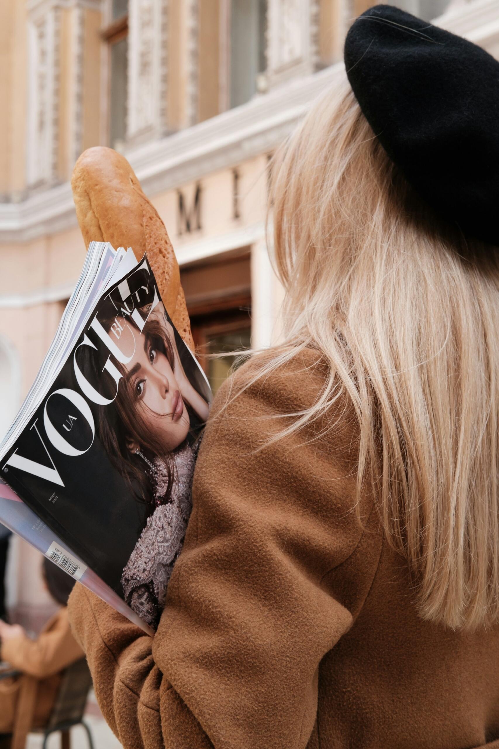 Osoba w brązowym płaszczu i czarnym beretem, trzymająca bagietkę i magazyn „Vogue”, na zewnątrz w pobliżu budynku.