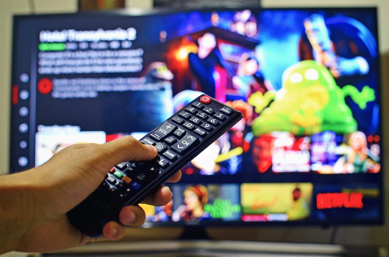 na pierwszym planie ręka trzymająca pilota, na drugim planie telewizor z włączonym netflixem