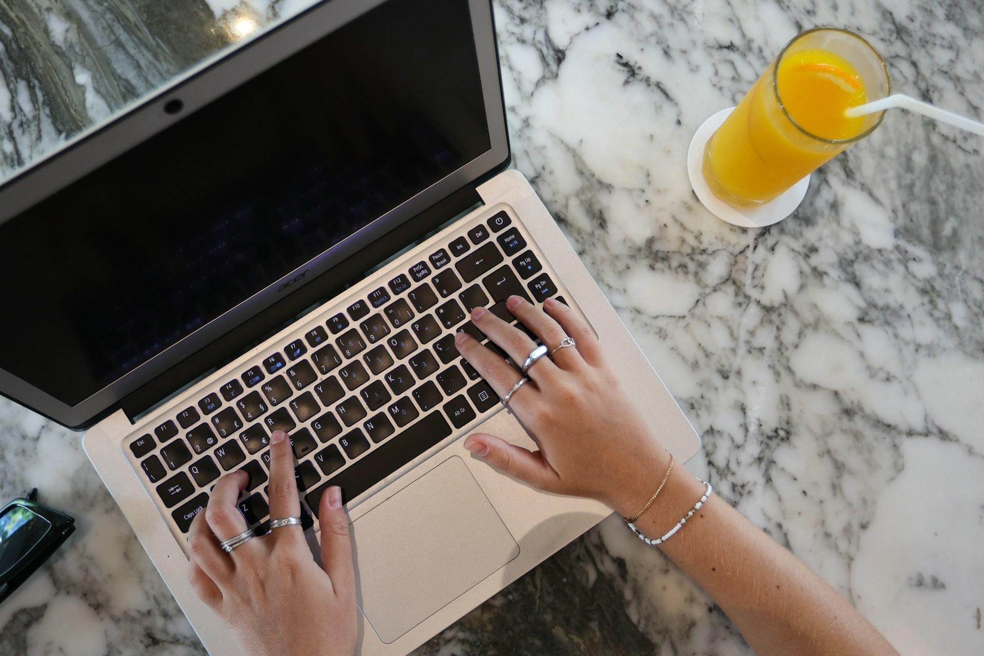 Laptop na marmurowym blacie, szklanka soku pomarańczowego, dłonie na klawiaturze.