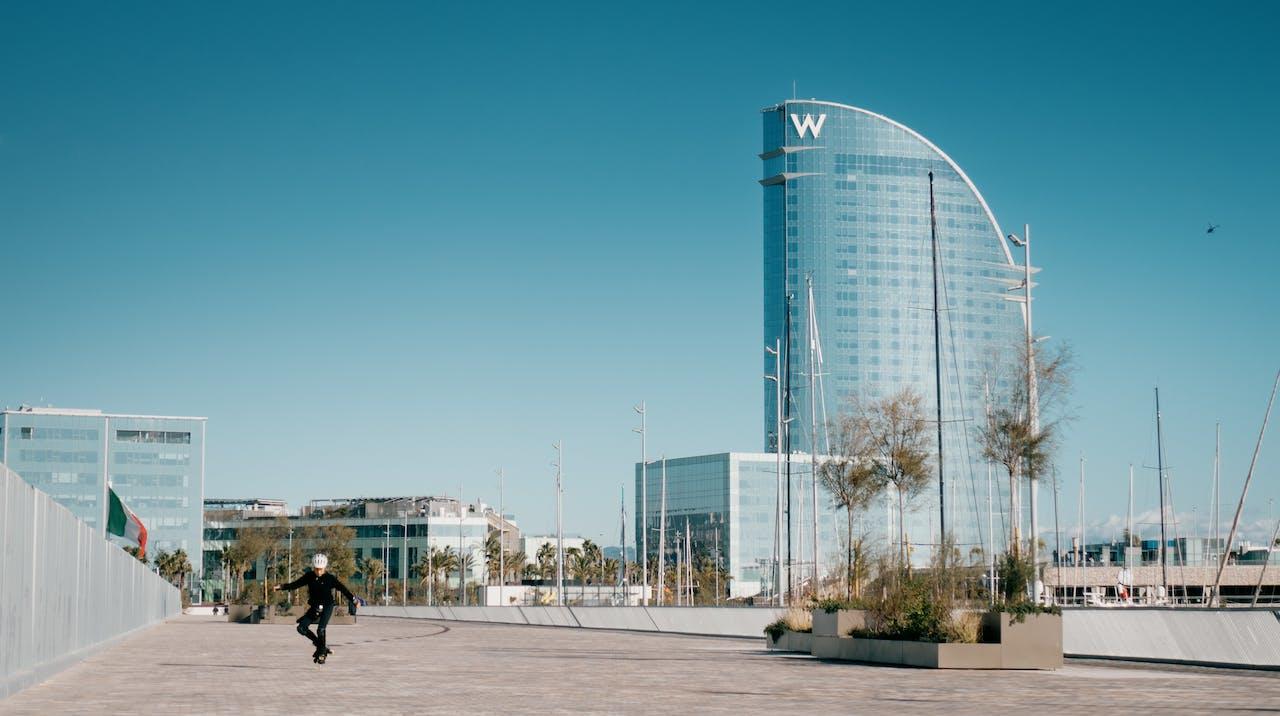 W hotel w tle, kilka nowoczesnych budynków, lampy uliczne, chlopak ubrany na czarno i skatujący na desce