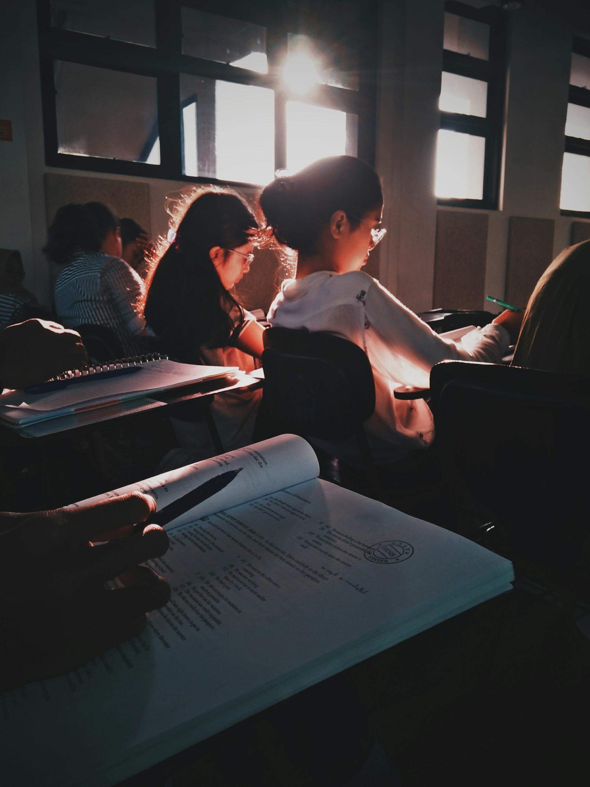 na pierwszym planie otwarta książka, w tle uczniowie, ciemna klasa, słońce przez okno