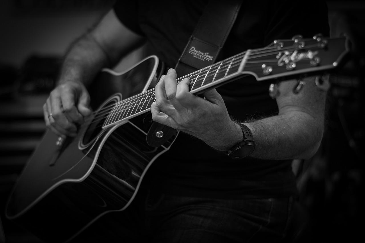 czarno białe zdjęcie, tułów mężczyzny grającego na gitarze, ciemna koszulka na krótki rękac, obrączka, zegarek