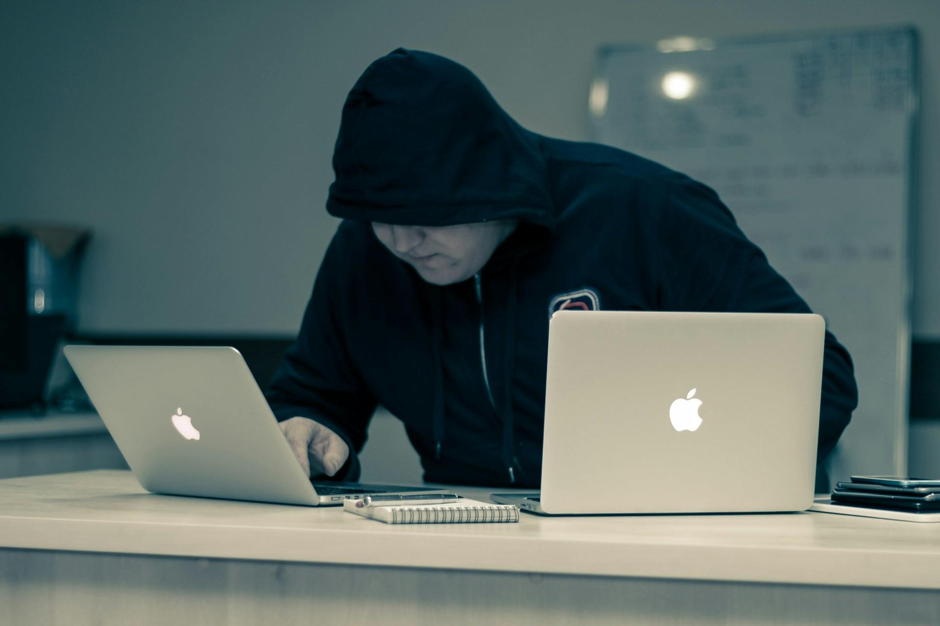 Mężczyzna w czarnej bluzie z kapturem naciągniętym na głowę pochylony nad dwoma laptopami marki Apple.