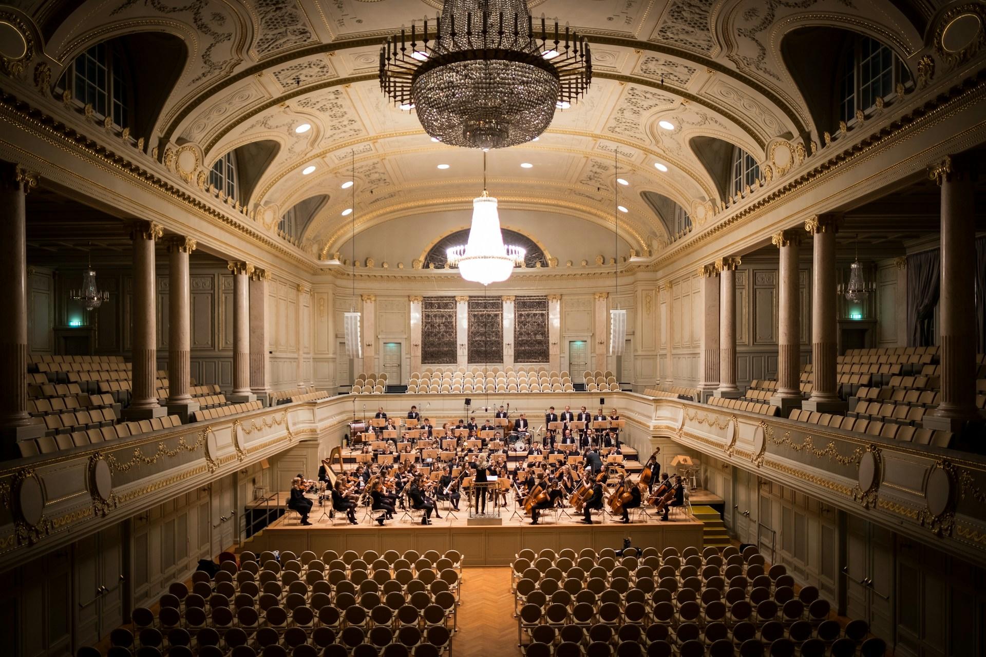 grająca orkiestra, wisząca lampa w cenutrym, puste siedzenia, kolumny, wystawny teatr