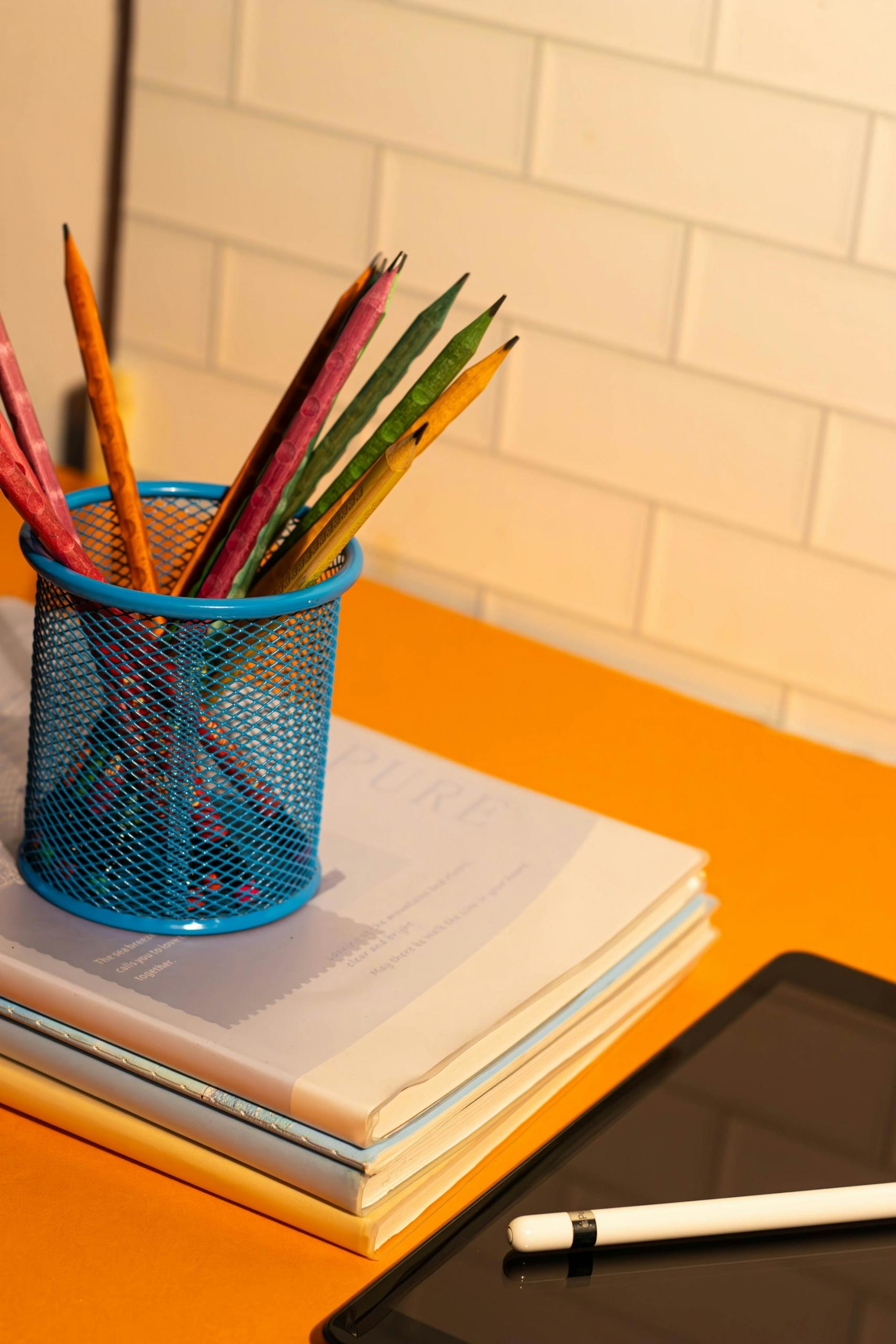 Niebieski pojemnik z ołówkami na książkach i laptop z długopisem na biurku.