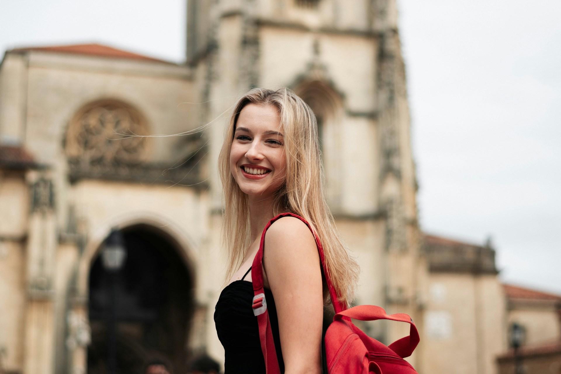 Uśmiechnięta blondynka z czerwonym plecakiem przy zabytkowym budynku.