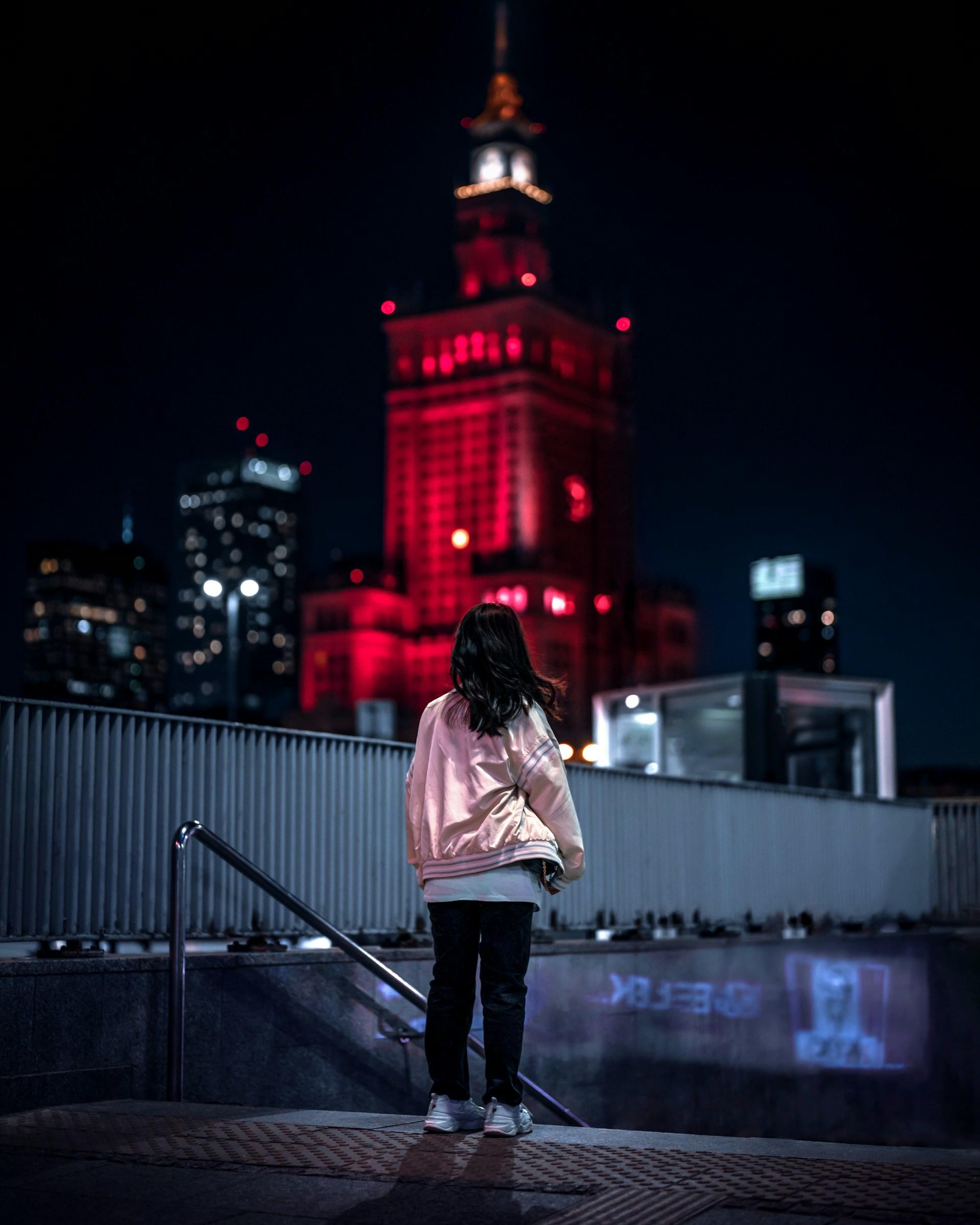 Warszawa, dziewczyna stojąca przed Pałacem Kultury oświetlonym na czerwono nocą.