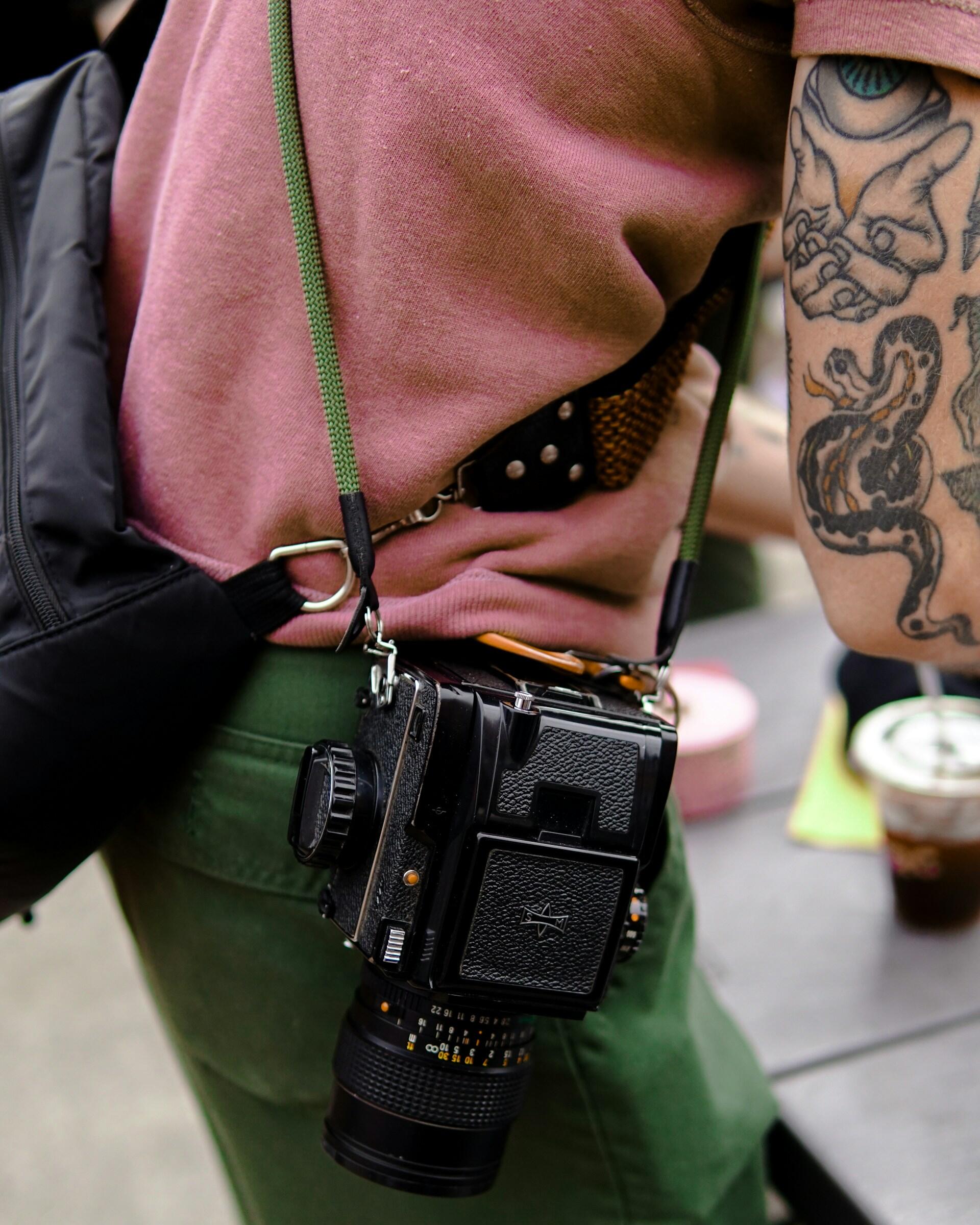 Fotograf z tatuażem w różowej koszulce z torbą i aparatem fotograficznym zawieszonym na szelce fotograficznej.