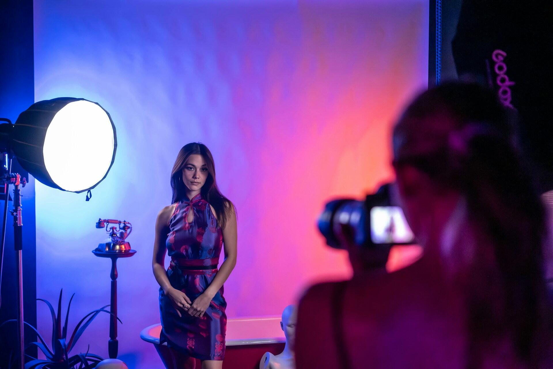 Fotograf na sesji z modelką w sukience obok telefonu przy kolorowym oświetleniu.